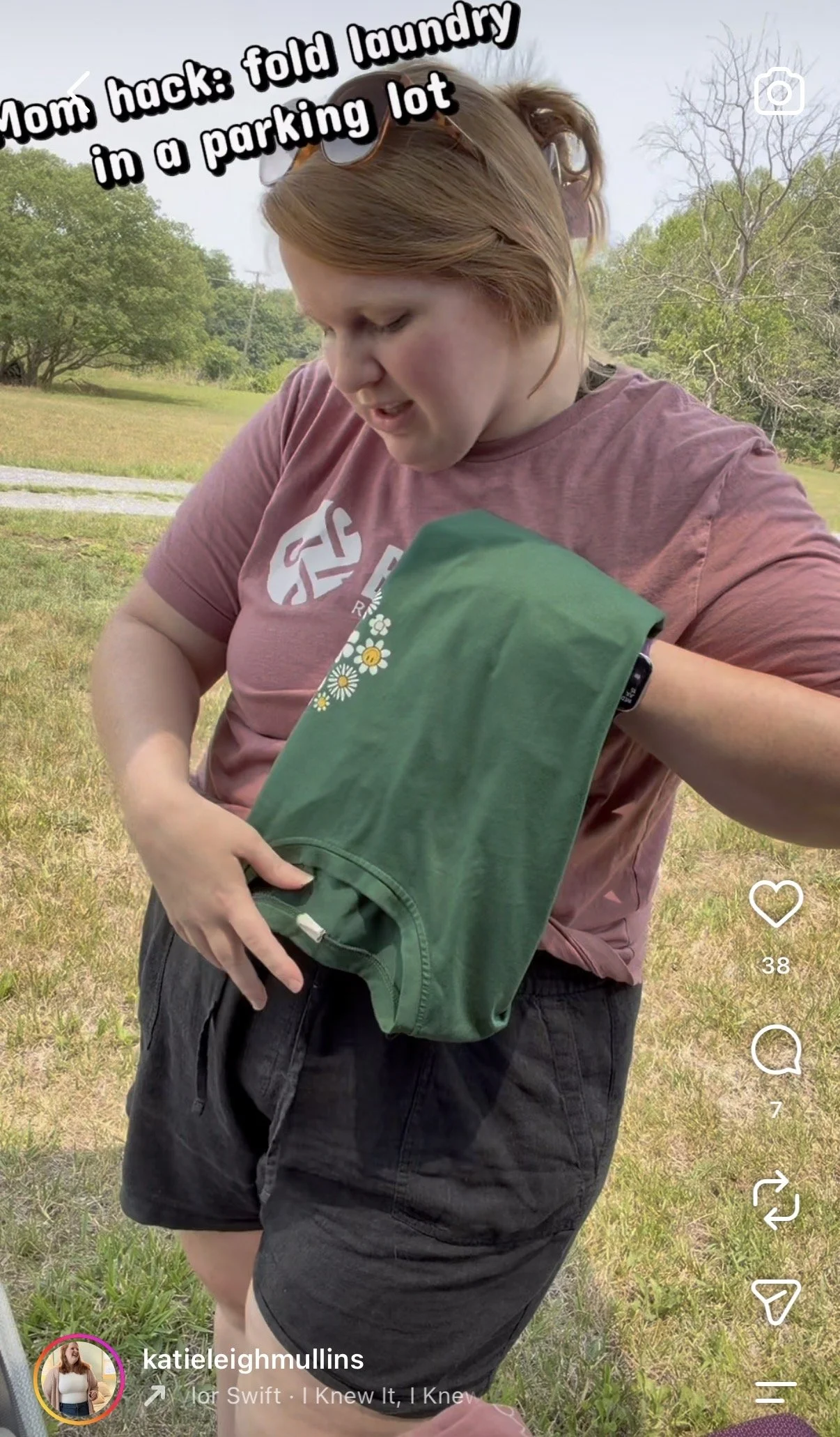 A young person with red hair and glasses on their head is holding a green item, possibly a folded piece of clothing or fabric, outdoors in a grassy area with trees in the background.