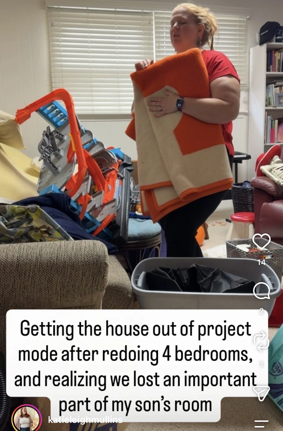 A woman in a red shirt holding folded blankets while standing in a cluttered living room with toys, furniture, and books, as she prepares to clear out her son's room.