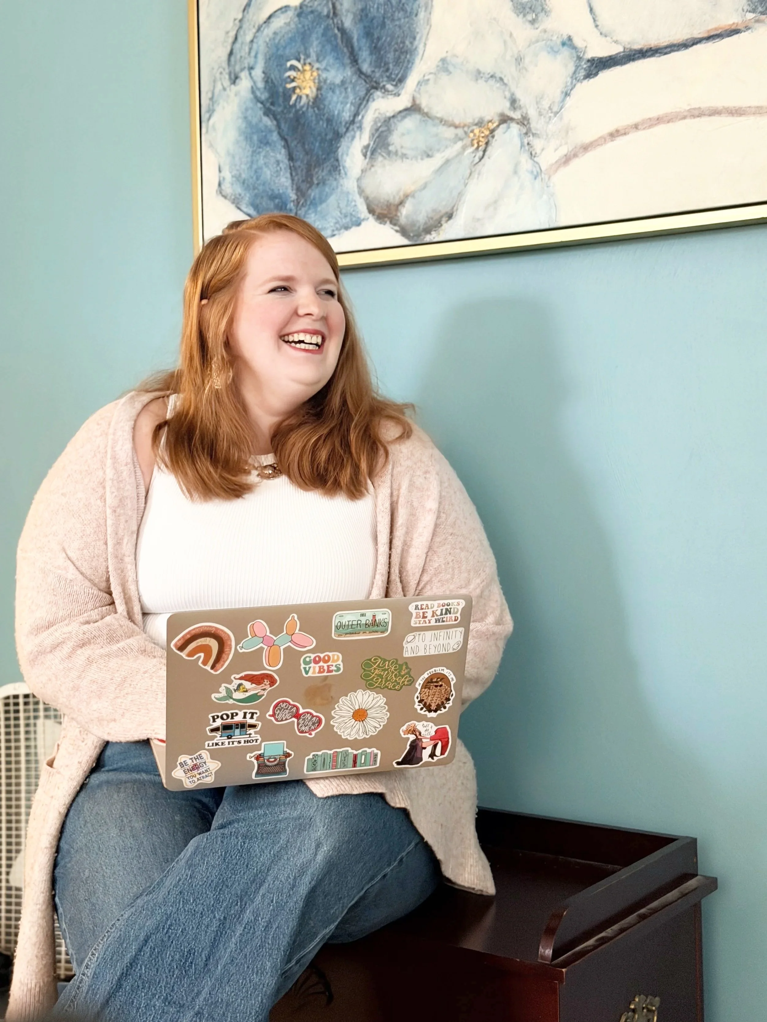A woman with red hair sitting on a bench, smiling at a laptop covered with colorful stickers, in a room with a teal wall and a framed abstract painting.