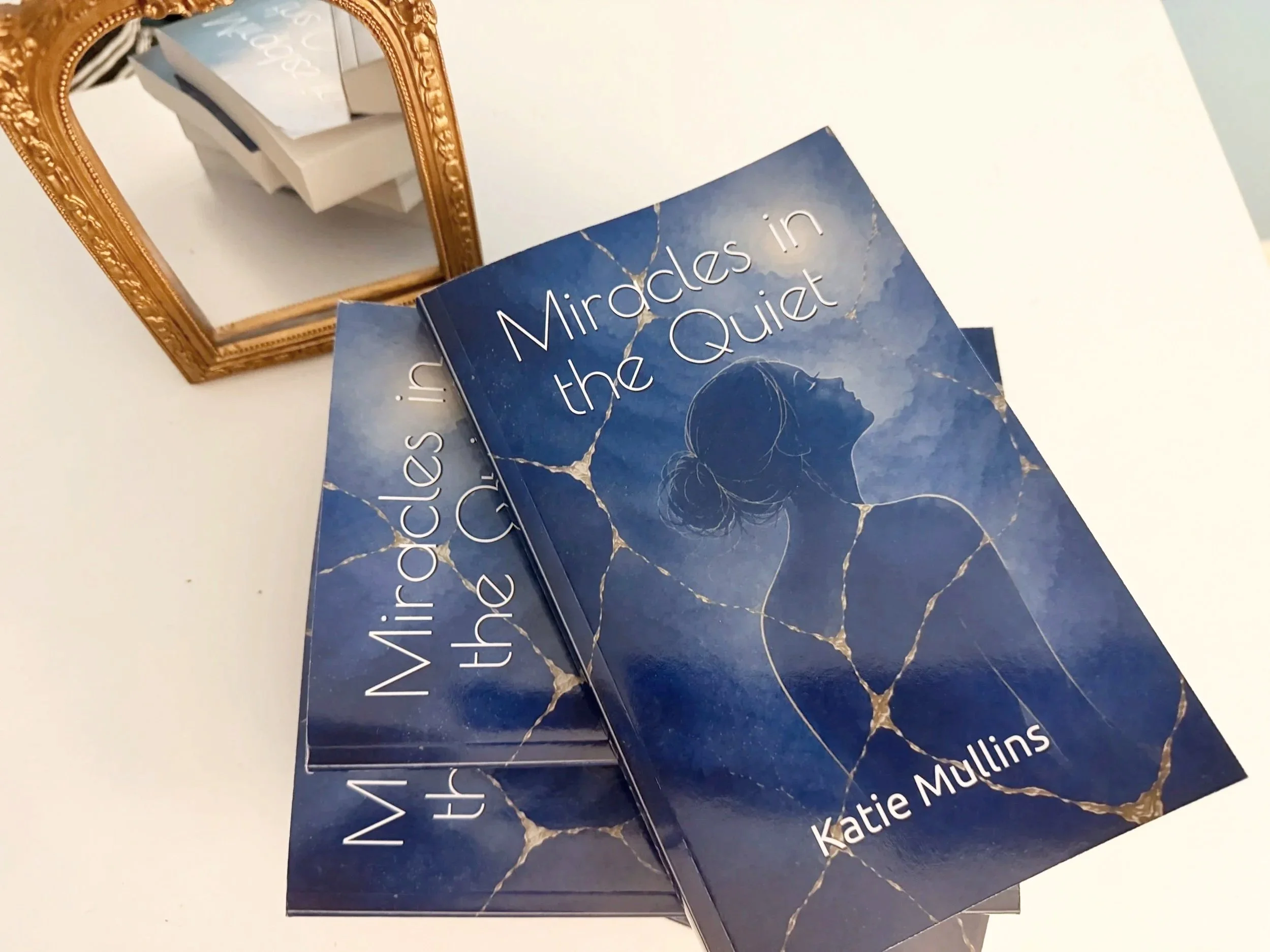 Close-up of multiple copies of a book titled "Miracles in the Quiet" by Katie Mullins, with a gold decorative mirror in the background on a white surface.