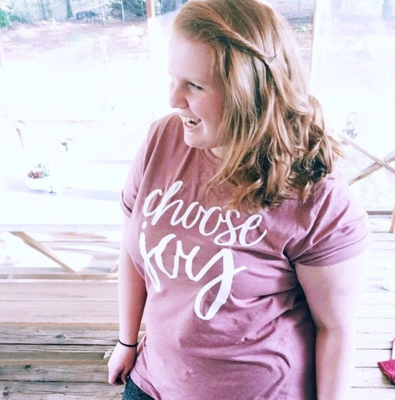 A woman with long red hair wearing a pink T-shirt that says 'choose joy'
