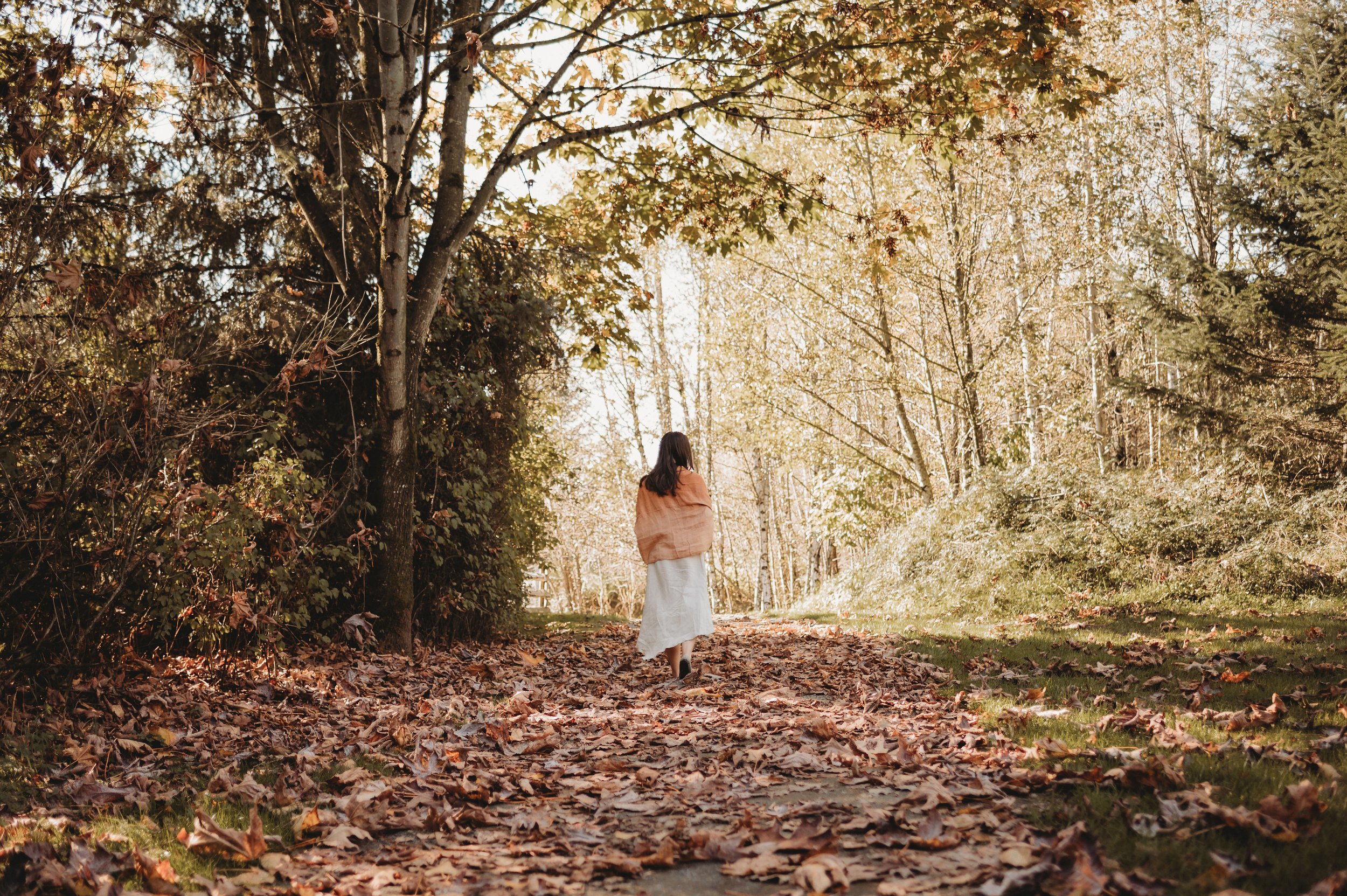A woman walking down a leaf-covered trail in a wooded area during autumn, surrounded by trees with changing leaves.
