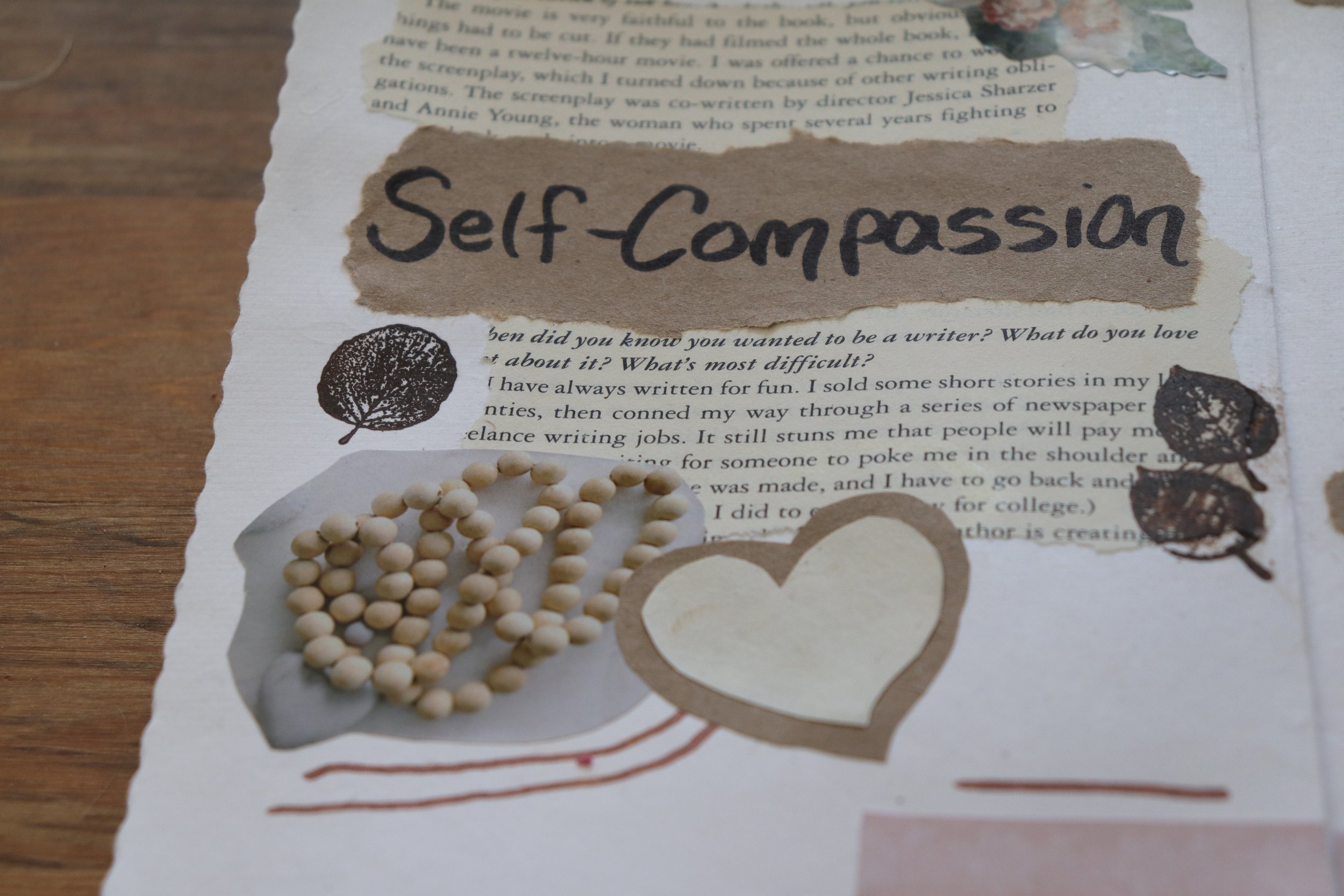A collage of paper pieces with the phrase 'Self-Compassion' handwritten on a brown paper strip. The collage includes various tear-edged papers with printed text, illustrations, and decorative elements like small beads and a tree symbol.