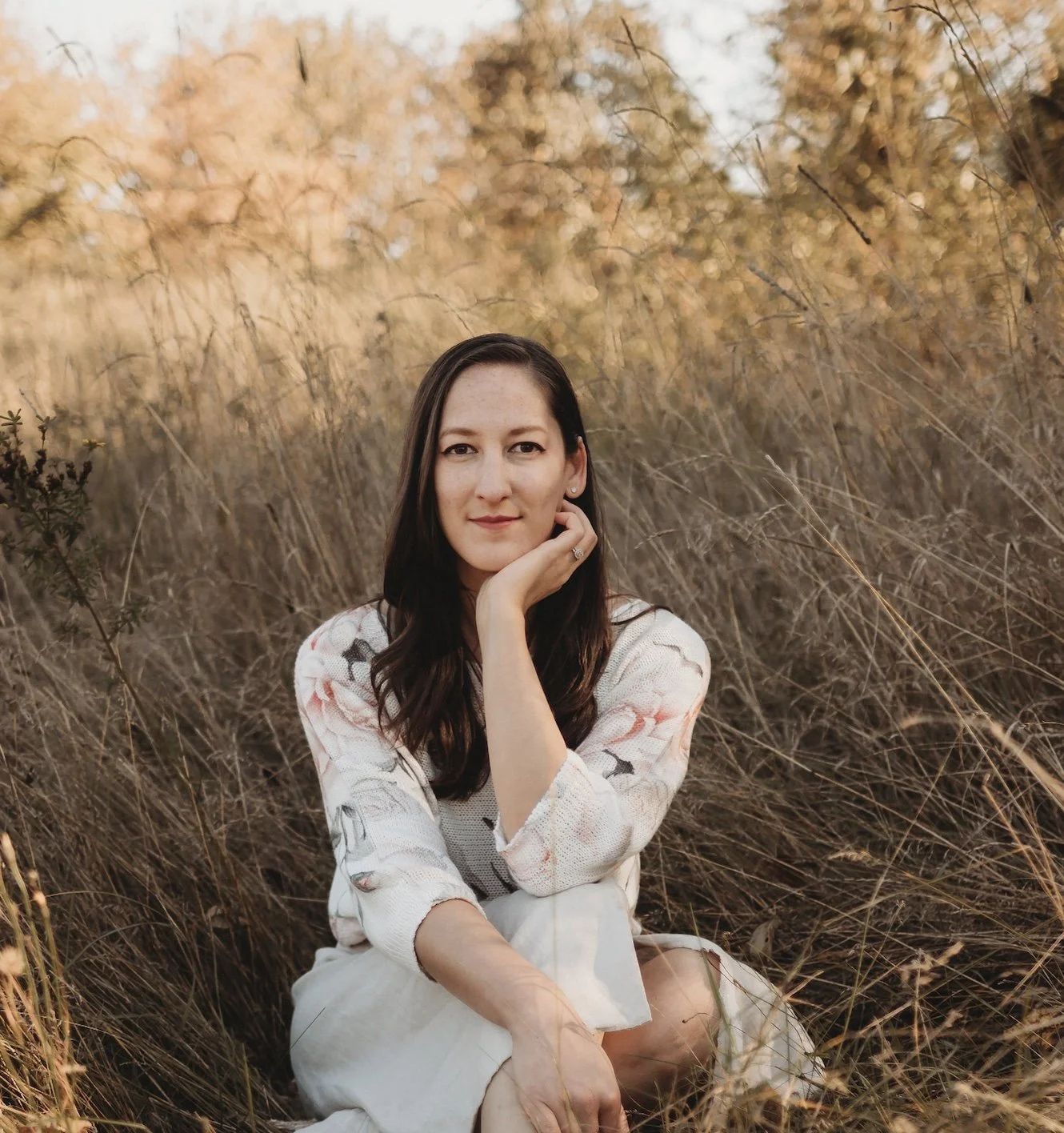 A woman with long dark hair sitting in tall, dry grass during sunset in a natural outdoor setting.