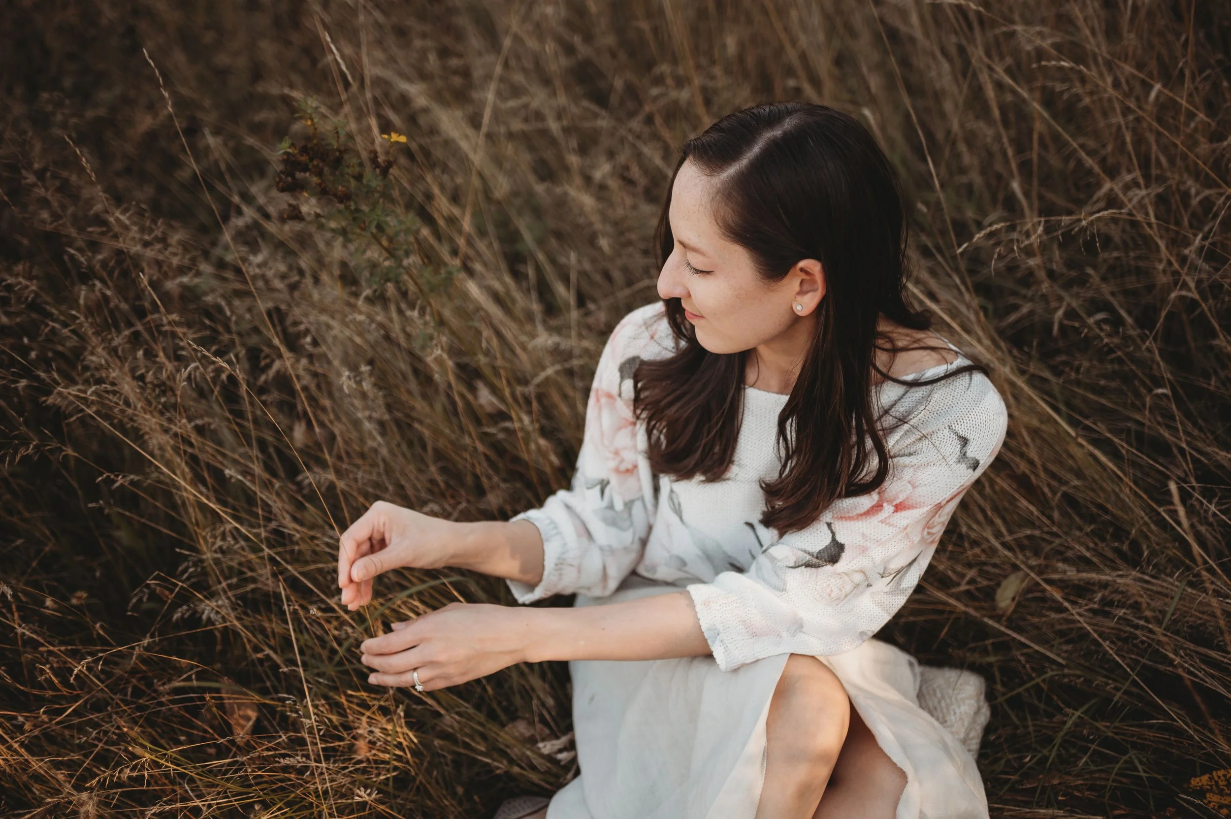 A woman with dark hair and earrings sitting in a field of dry grass, with her eyes closed and a slight smile, wearing a white top with floral patterns.