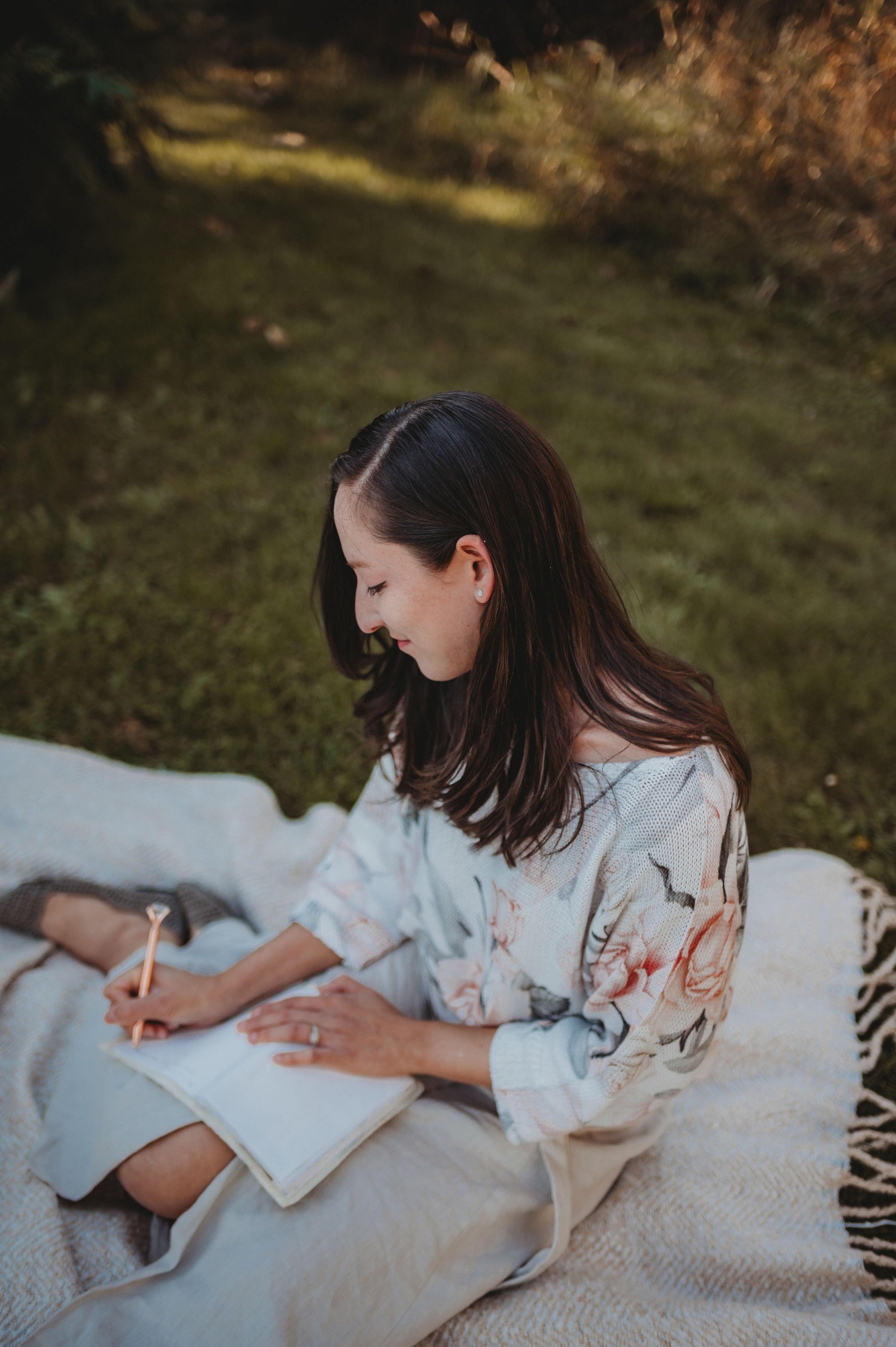 A woman with long dark hair and earrings, wearing a floral robe, sitting on a bed outside on a blanket, writing in a notebook with a pen, with greenery in the background.