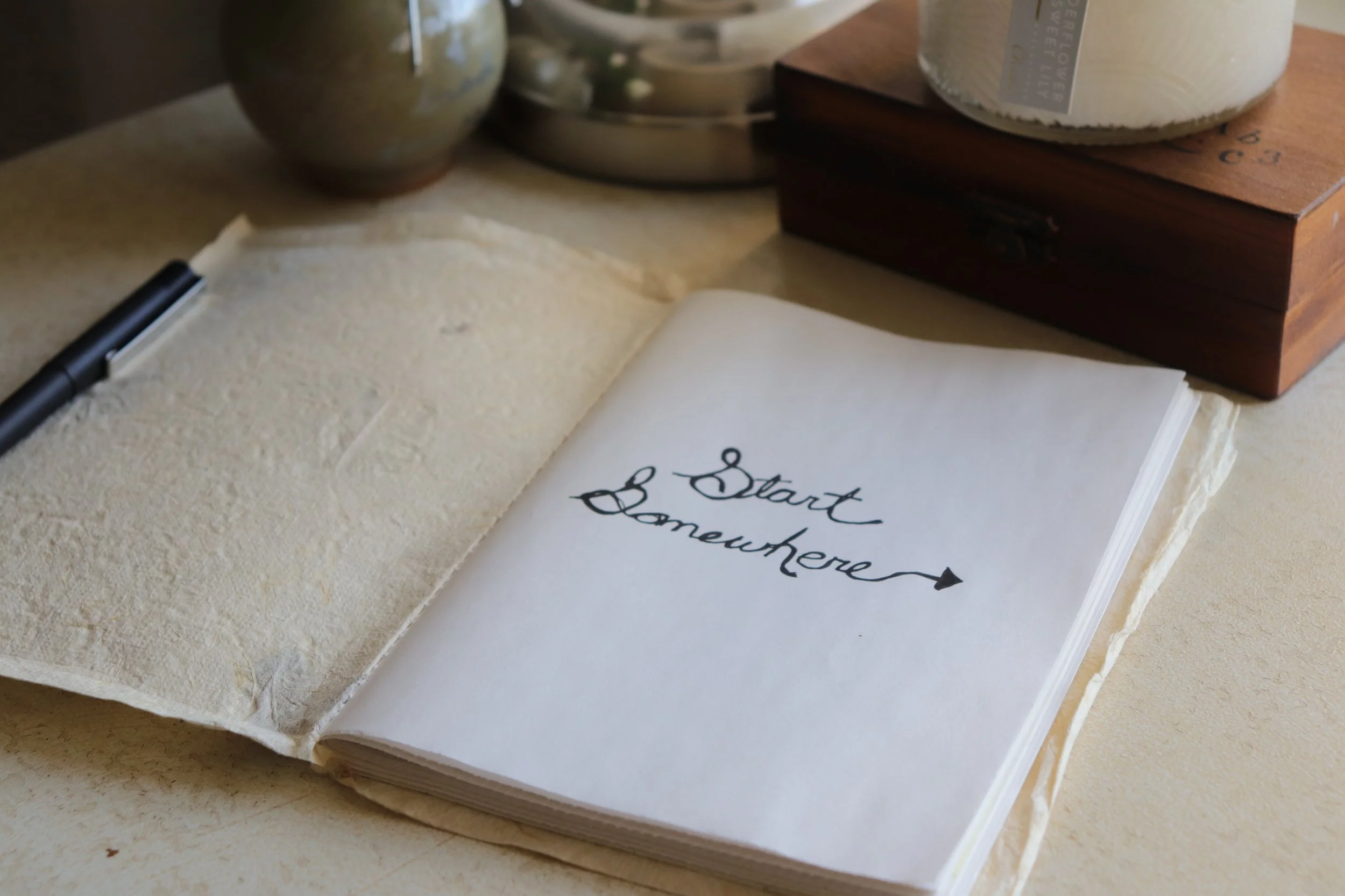 An open guest book with the words 'Start Somewhere' written on the page. A black pen lies on the pages, and in the background, there are ceramic and wooden decorative items.
