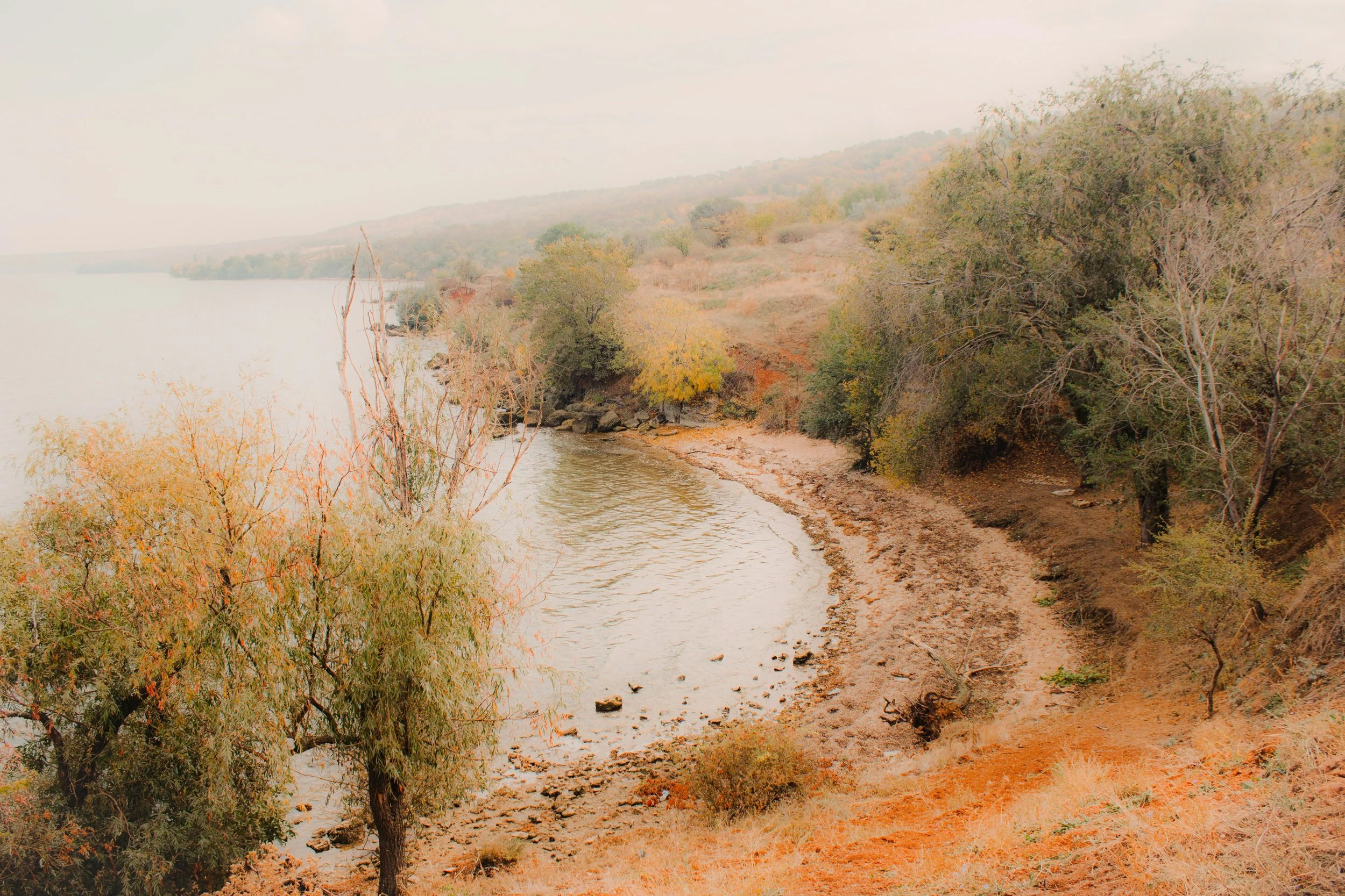 A river flowing alongside a narrow dirt trail with trees and shrubs on both sides, leading toward distant hills under an overcast sky.