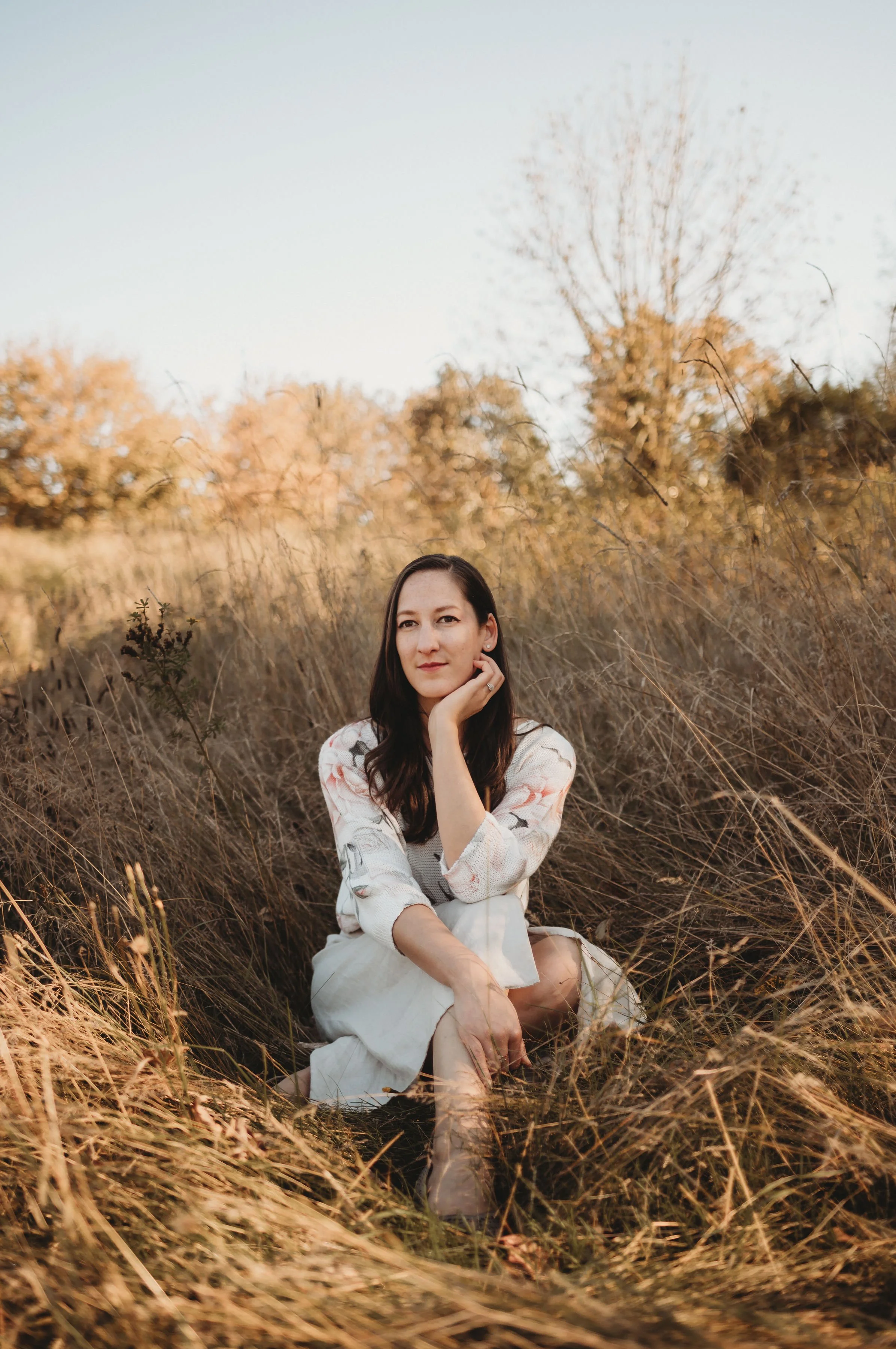 A woman with long dark hair sitting in a field of dry grass and trees, wearing a white and pink floral blouse and light-colored pants.
