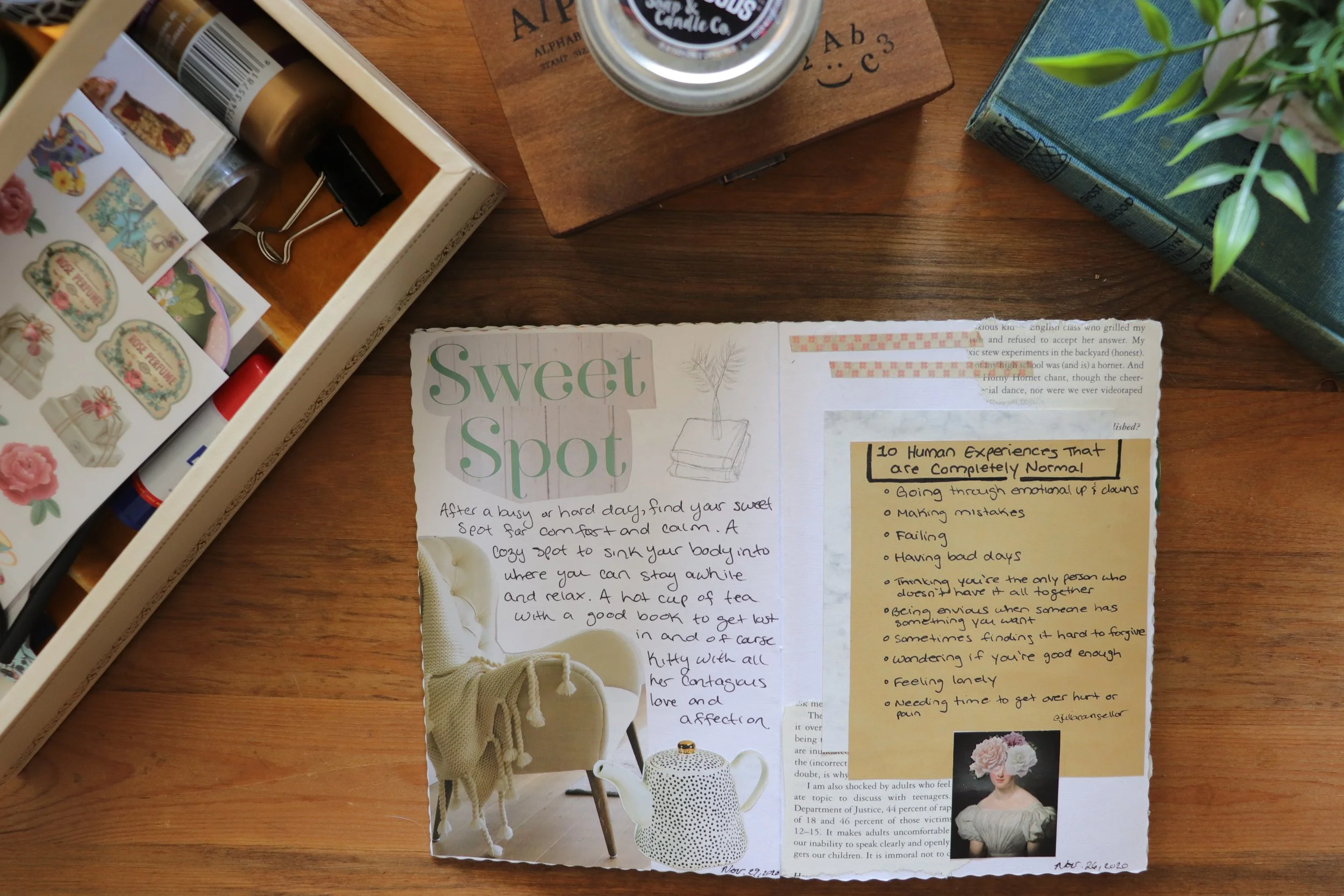 Flat lay of various items on a wooden surface, including a jar with a black label, a small cutout paper with handwritten notes about human experiences and a cozy spot for relaxation, a blue book, a green plant, and a box of assorted decorative stickers with floral and bow designs.