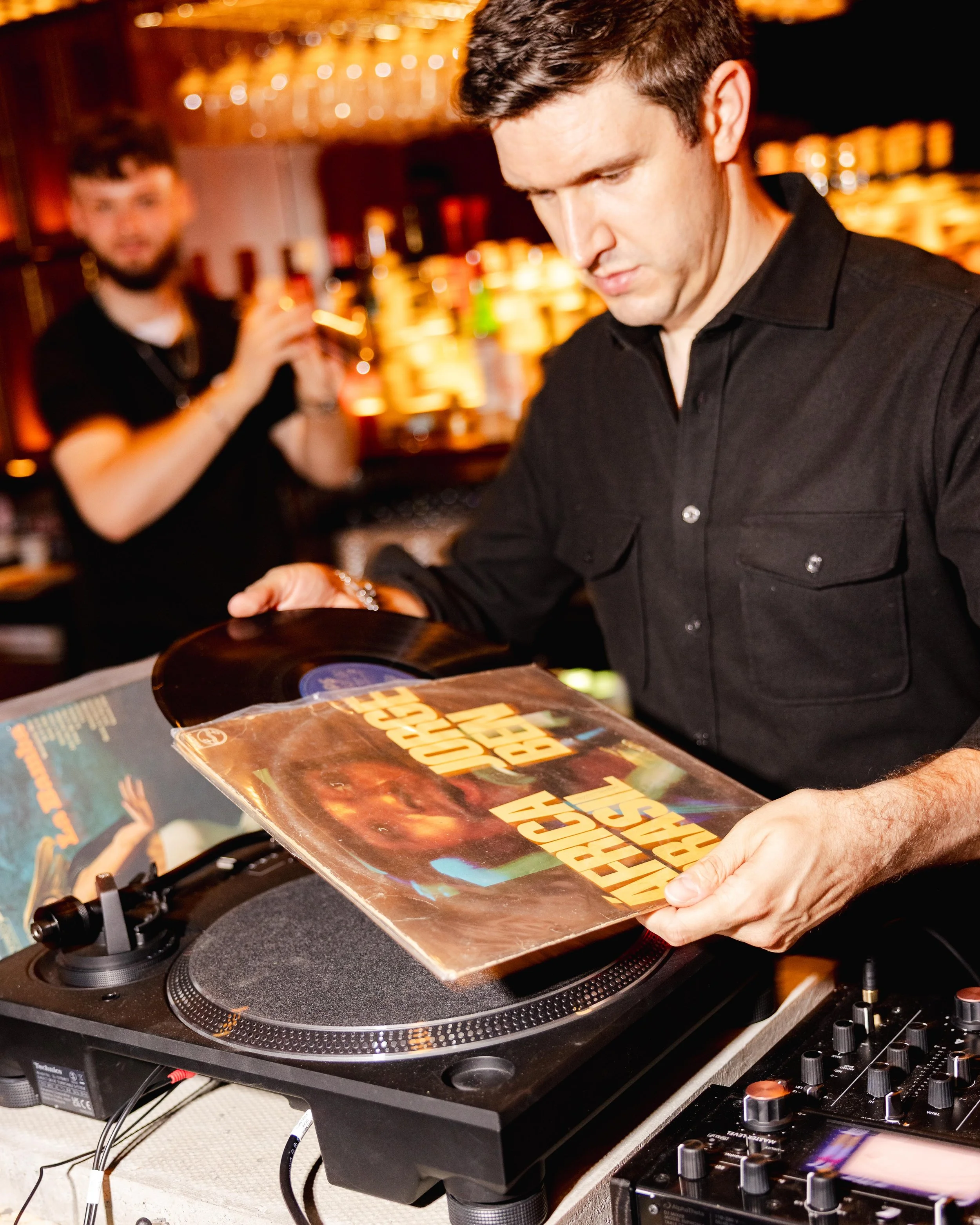A DJ in a black shirt holding a vinyl record in front of a DJ turntable, with a blurry background of a bar with shelves of liquor bottles and glasses.