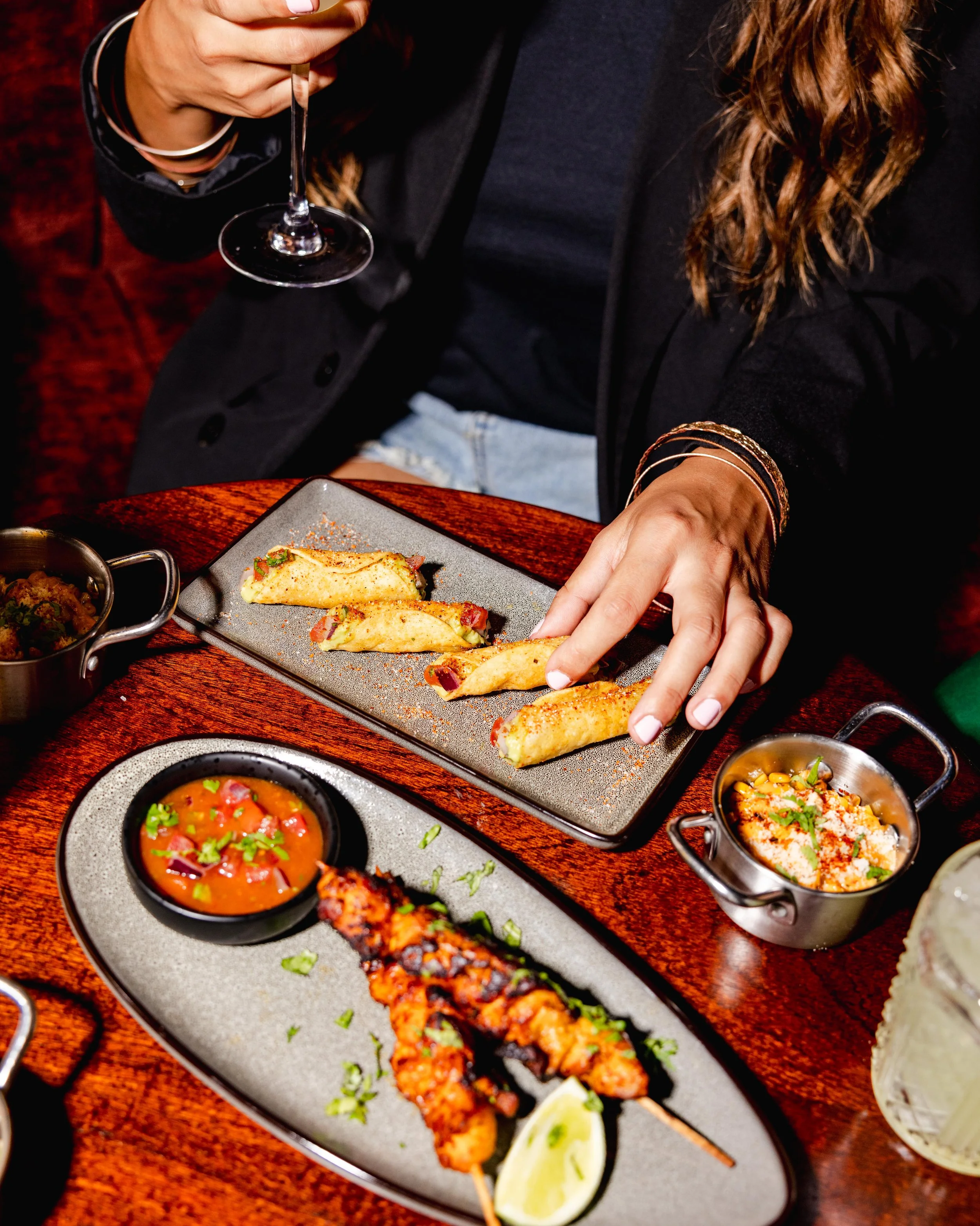 A person holding a wine glass, seated at a table with various Mexican dishes including tacos, dips, and sauces.