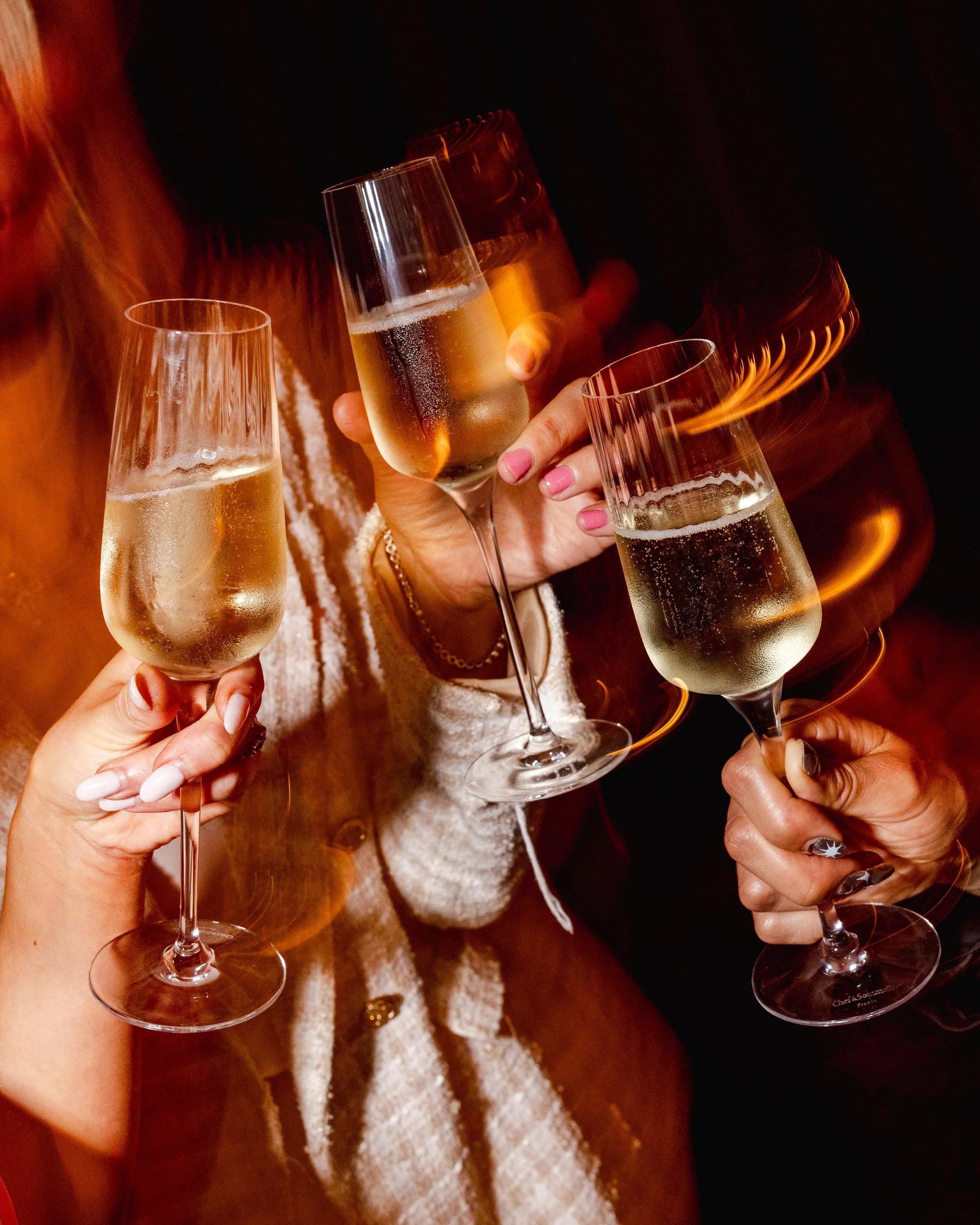Close-up of four women’s hands holding champagne glasses in a toast, with a dark background and one woman wearing a cream-colored textured coat.