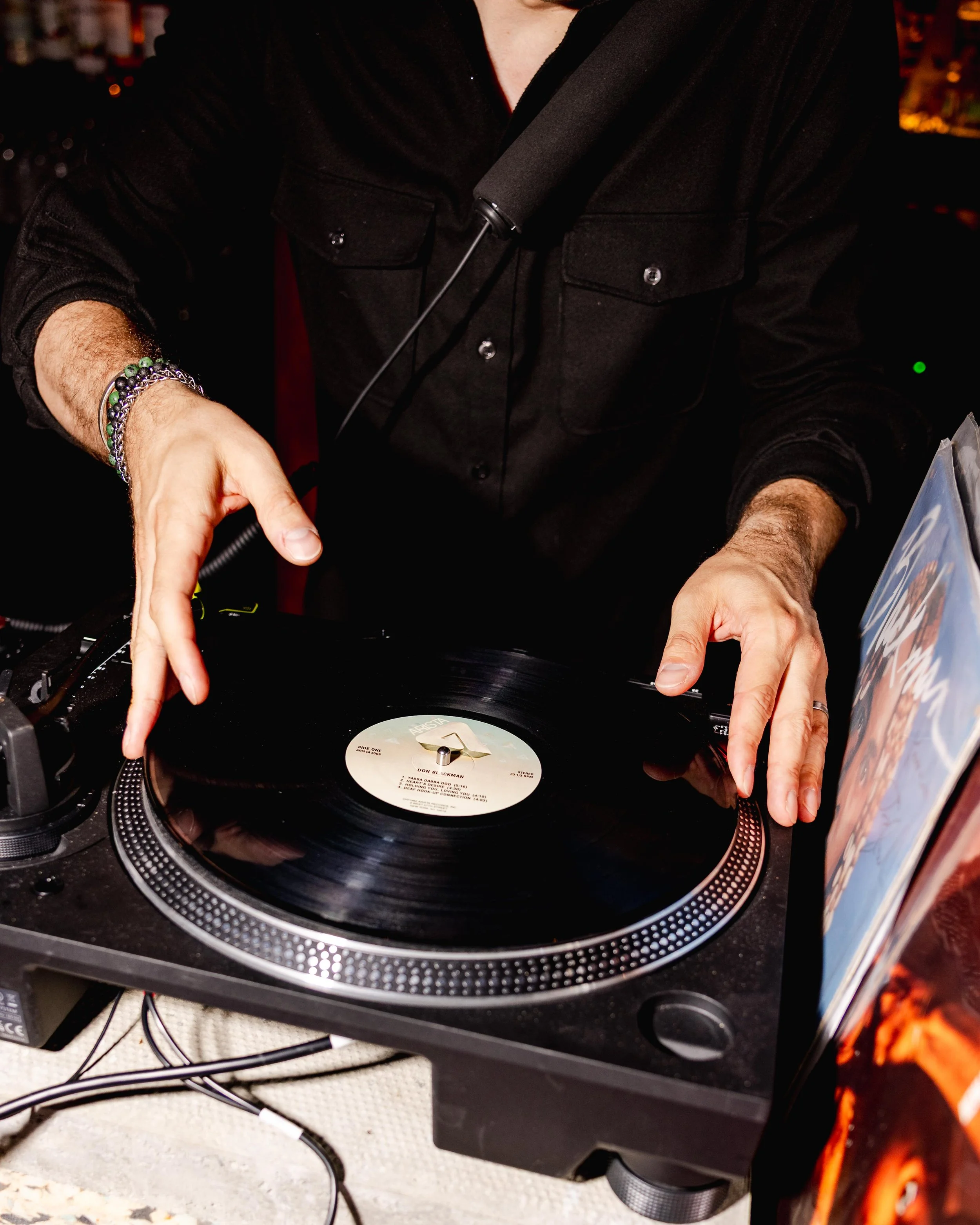 Person DJing with turntable and vinyl record, wearing black shirt and bracelets, in a dimly lit room.