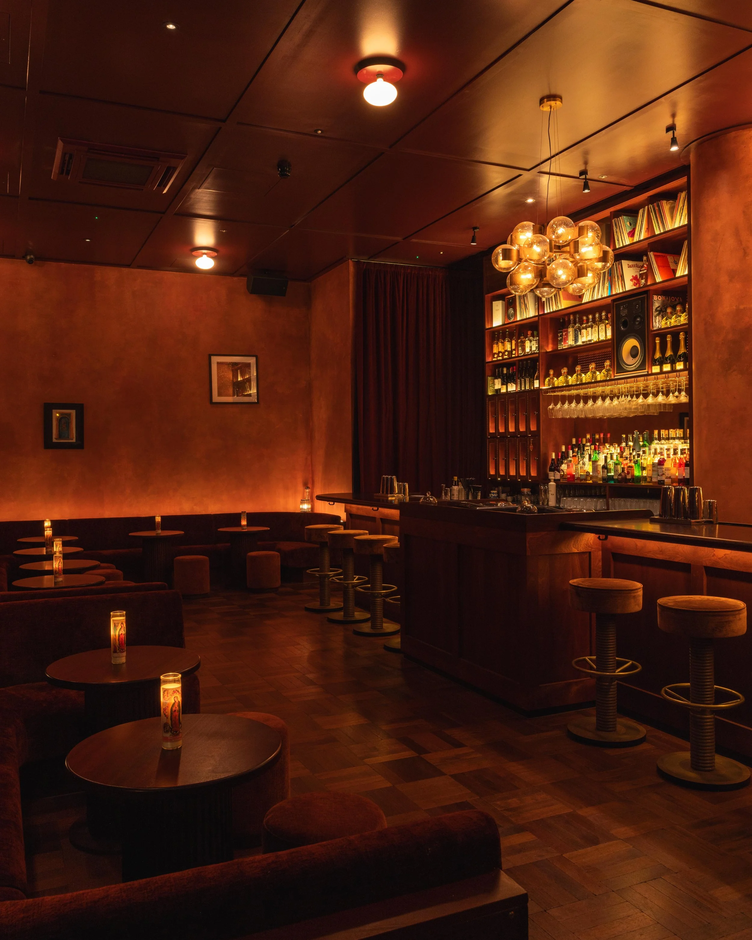 A dimly lit bar with wooden furniture, a well-stocked shelf of liquor bottles, and a modern chandelier hanging from the ceiling.