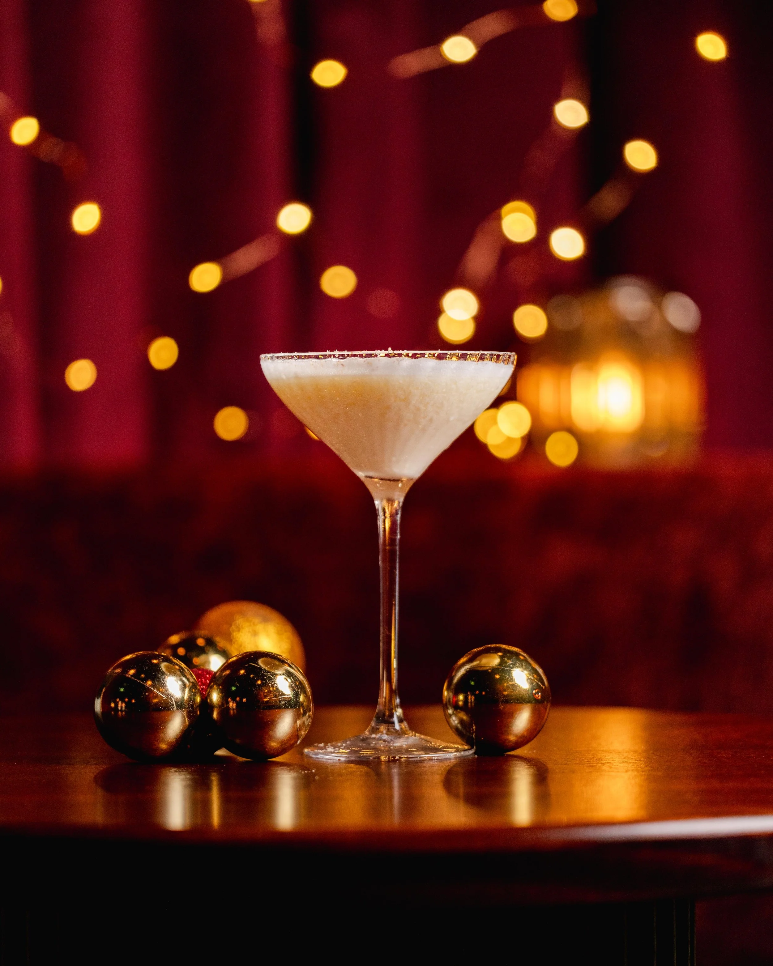 A cocktail glass with a salted rim filled with a frothy drink, surrounded by shiny gold Christmas ornaments, against a backdrop of red curtains and blurred yellow festive lights.