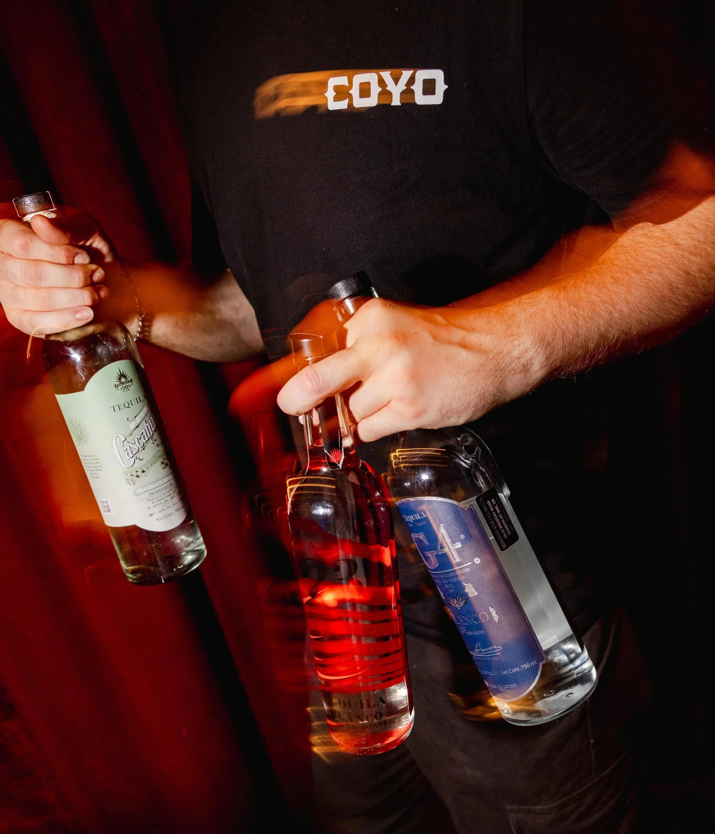 Close-up of a person wearing a black shirt with white text 'COYO' and holding a bottle of tequila, with several bottles of alcohol on a dark surface.