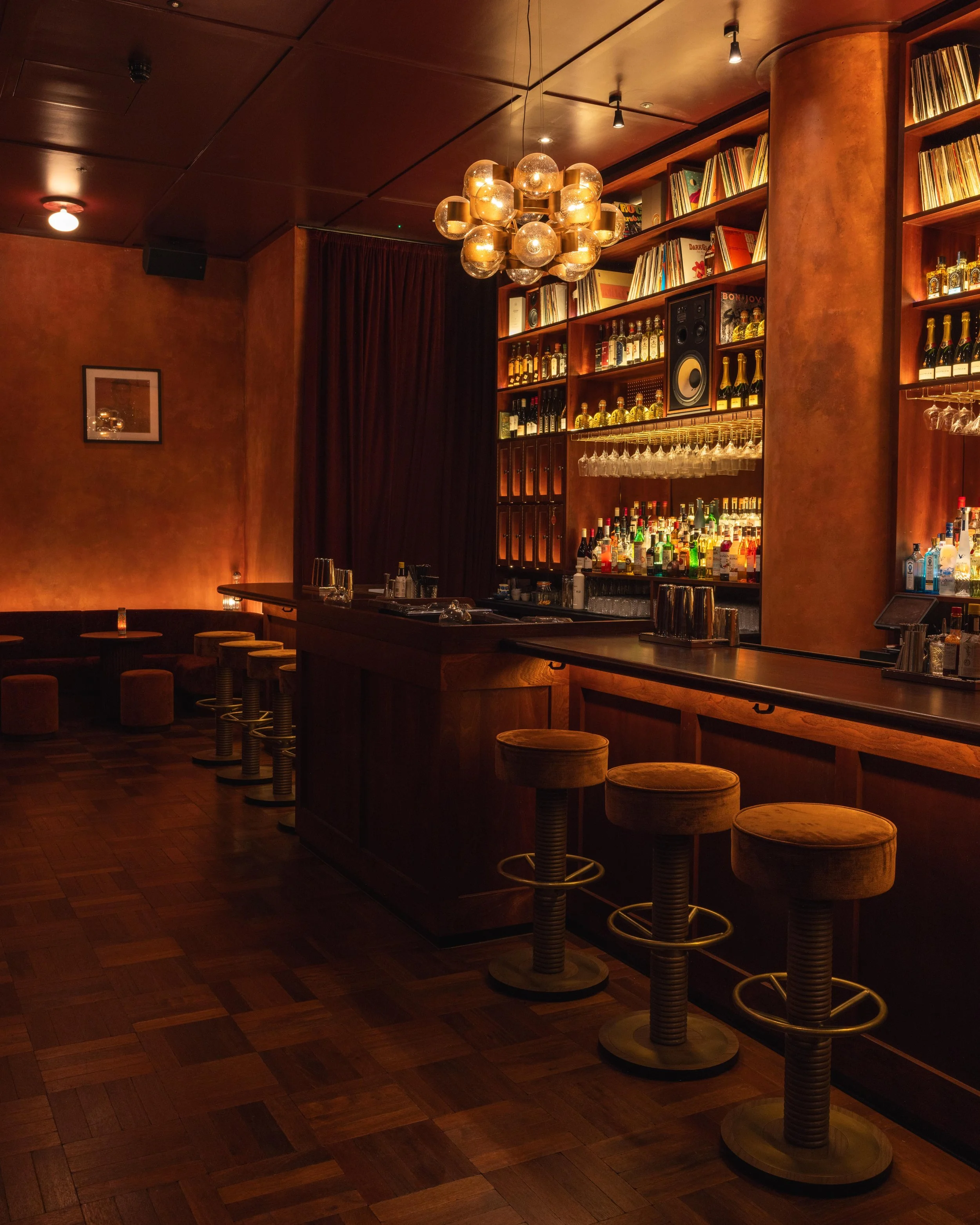 A dimly lit bar with a wooden countertop, several bar stools with cushioned seats, a large wooden shelf behind the bar stocked with liquor bottles and glassware, a decorative light fixture hanging from the ceiling, and framed artwork on the wall.