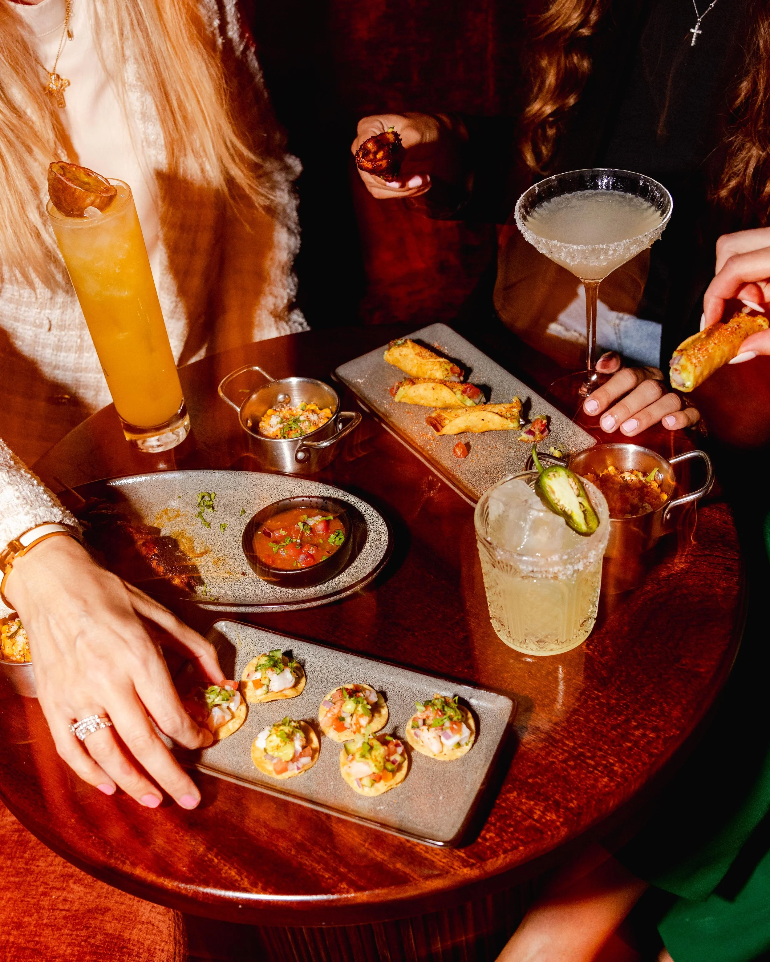 People at a restaurant table enjoying drinks and appetizers, including a yellow cocktail with a pineapple garnish, a margarita with a salted rim, and a plate of assorted mini tacos, with a focus on their hands and beverages.