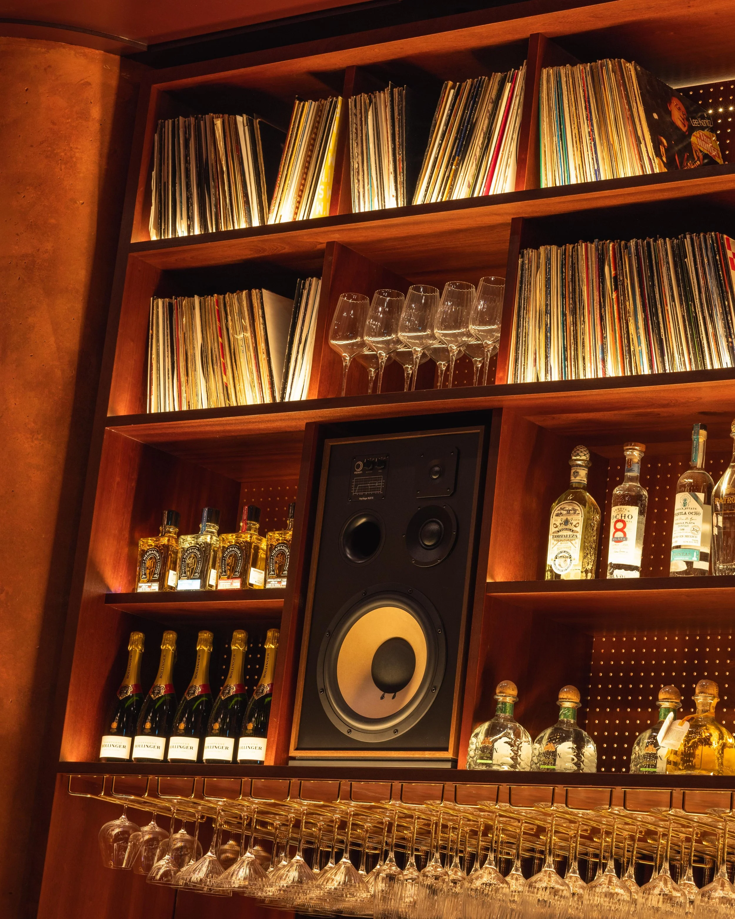 Wooden shelves holding vinyl records, glassware, liquor bottles, and a black speaker with large yellow and black speaker cone.