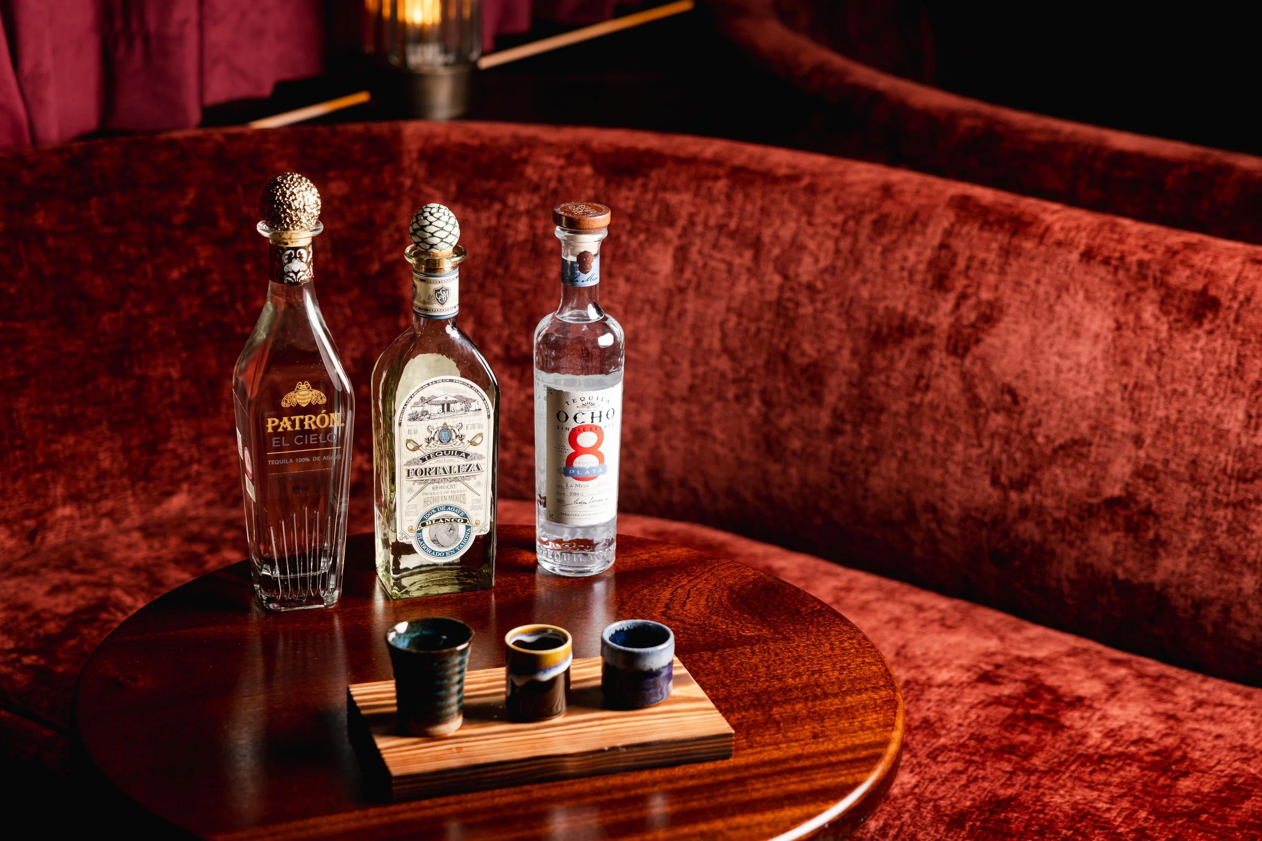 Three bottles of alcoholic beverages on a wooden table with sake cups in front, set against a plush red velvet background.