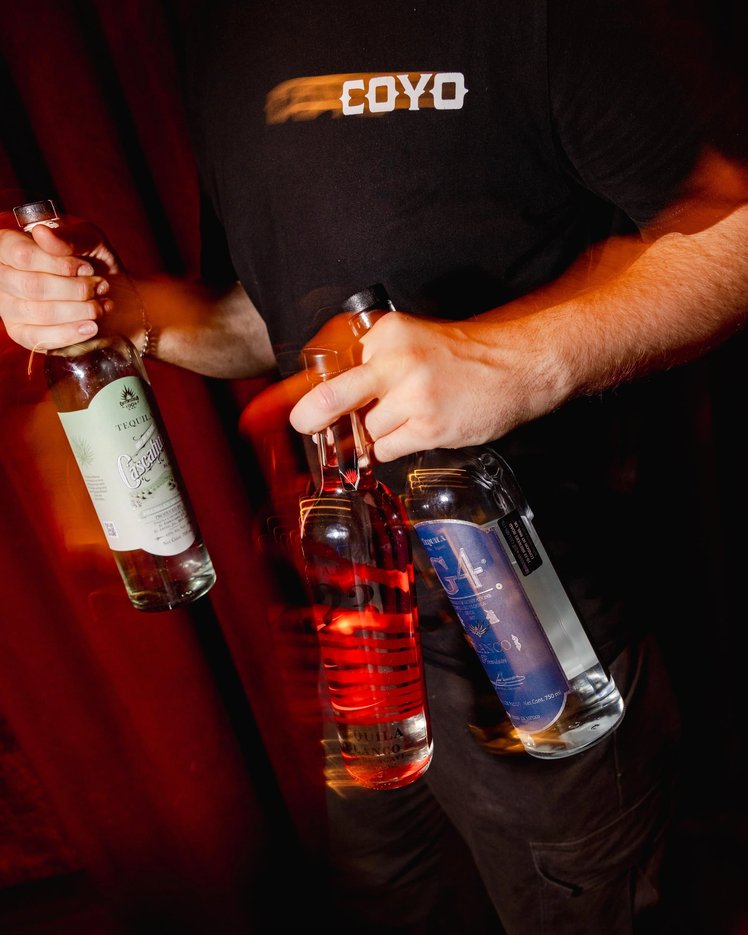 Person holding a bottle of tequila and a glass, wearing a black shirt with a logo on it.