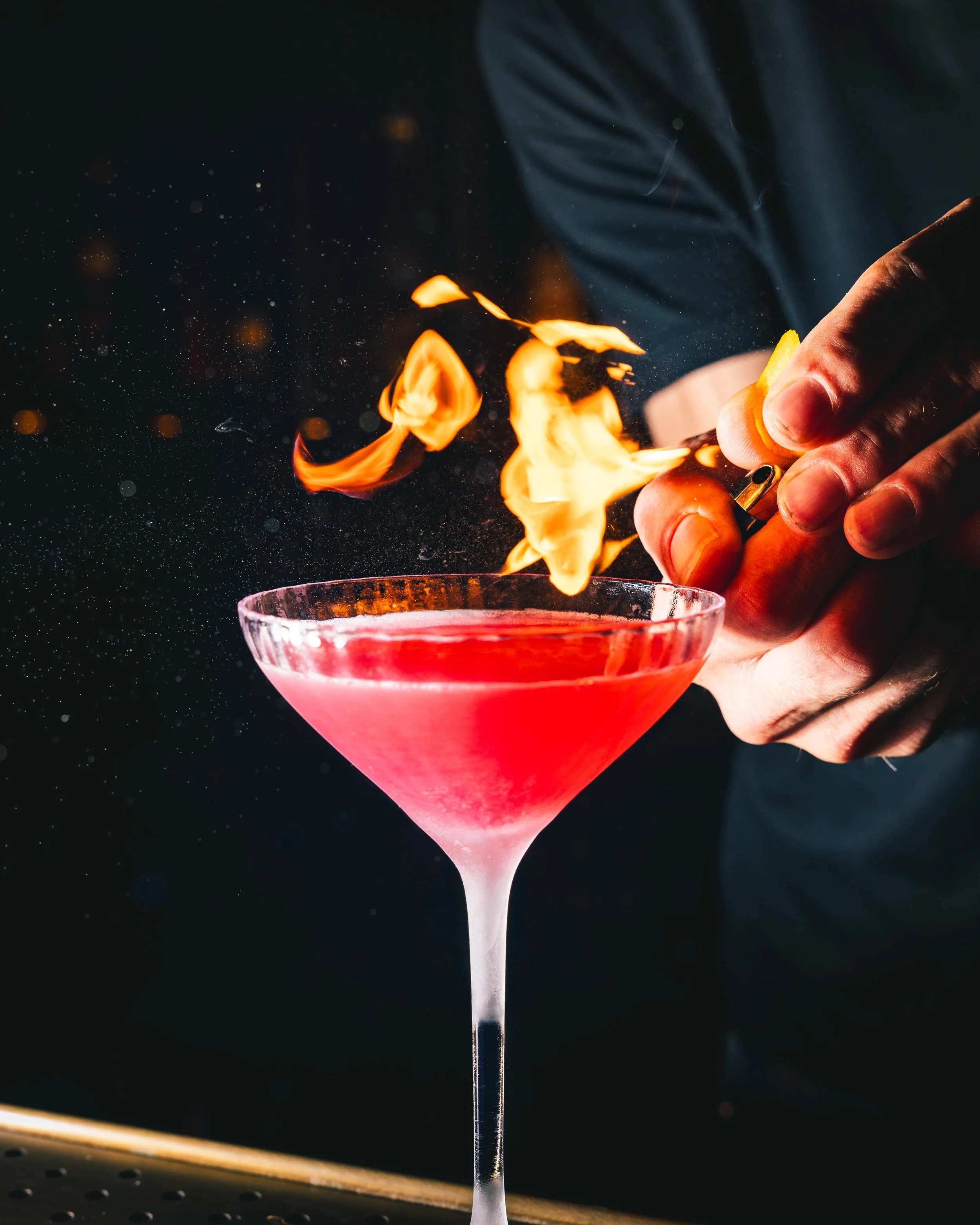 A close-up of a pink martini glass with a cocktail, being lit on fire by a flame held with fingers, against a dark background with tiny floating particles.