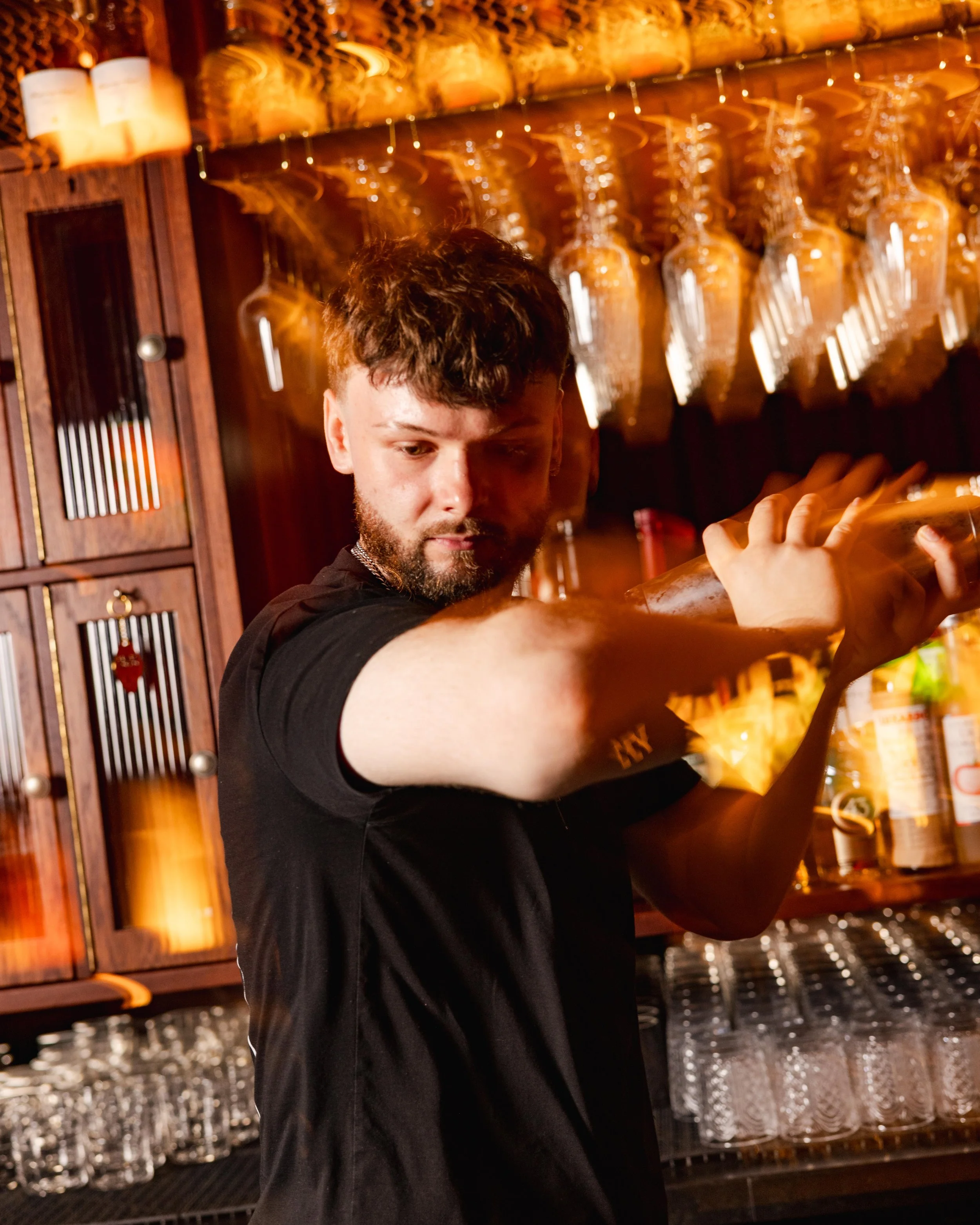 A bartender with short brown hair and a beard pouring a drink at a bar with a background of hanging wine glasses and bottles.