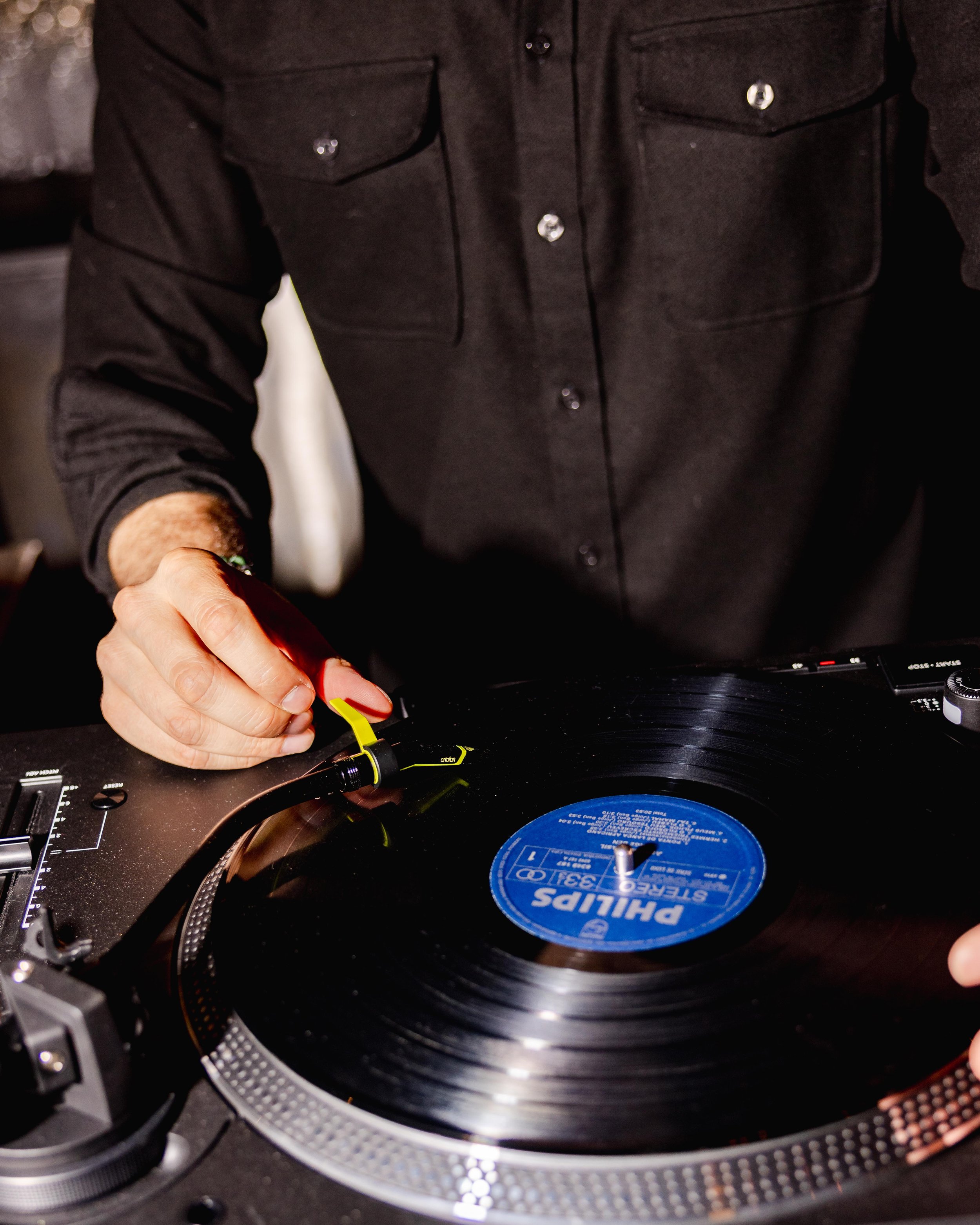 Close-up of a person's hand adjusting the tonearm of a black vinyl record turntable, with a blue record label visible on the spinning record.