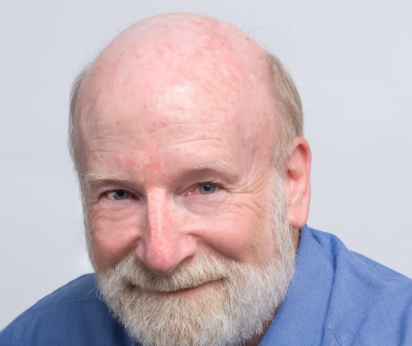 Close-up headshot portrait of William leaning toward the camera and smiling, wearing a blue shirt