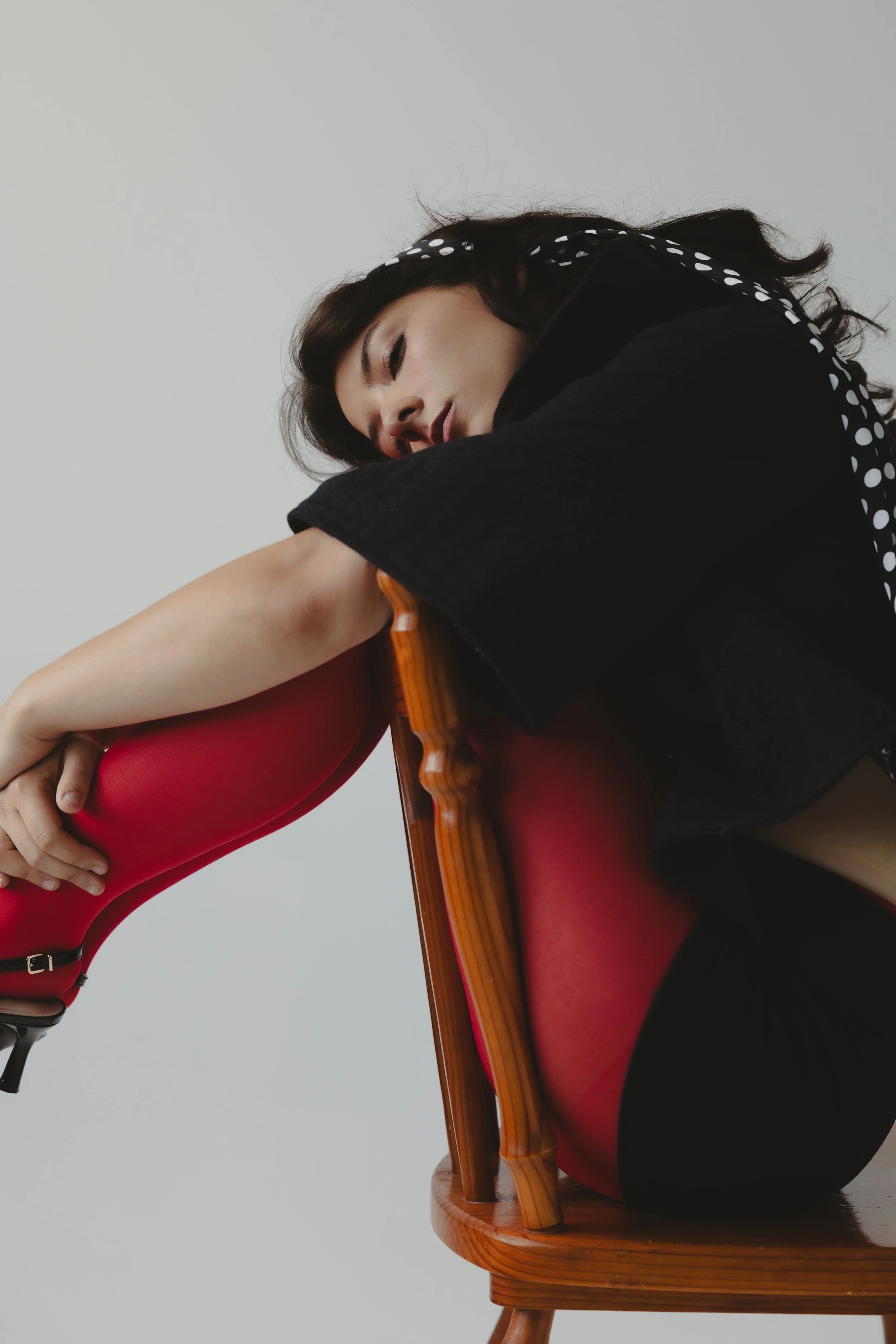 A woman with dark hair leaning on a wooden chair with a glossy finish. She is wearing bright red pants and a black jacket with a dotted pattern on the sleeve. The background is plain and neutral.