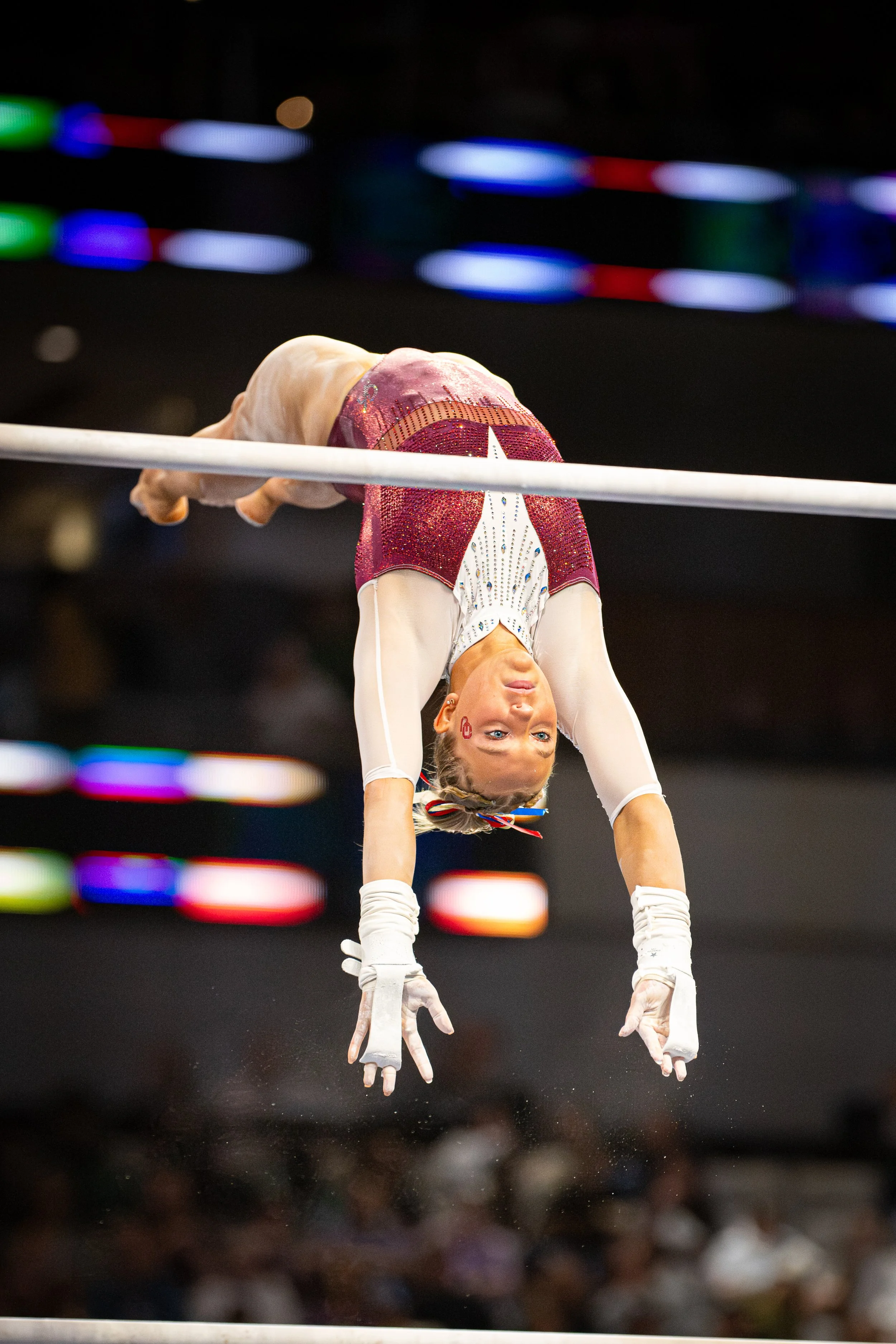 A female gymnast performing on the uneven bars during a competition, suspended upside down with her hands gripping the bar and her body extended