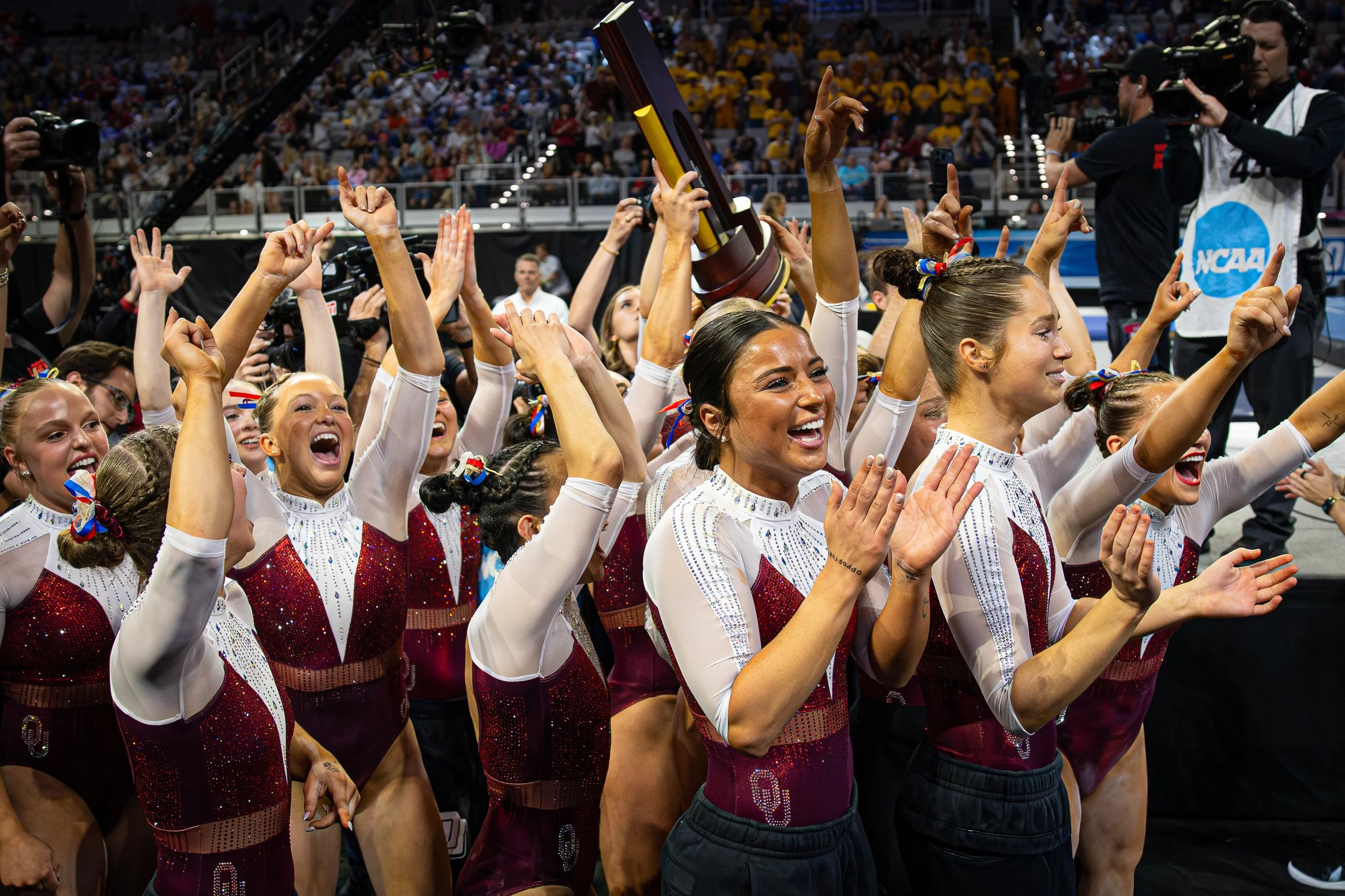 Women athletes in red and white uniforms celebrating with team members and holding a trophy at an indoor sports arena, with a crowd of spectators in the background.