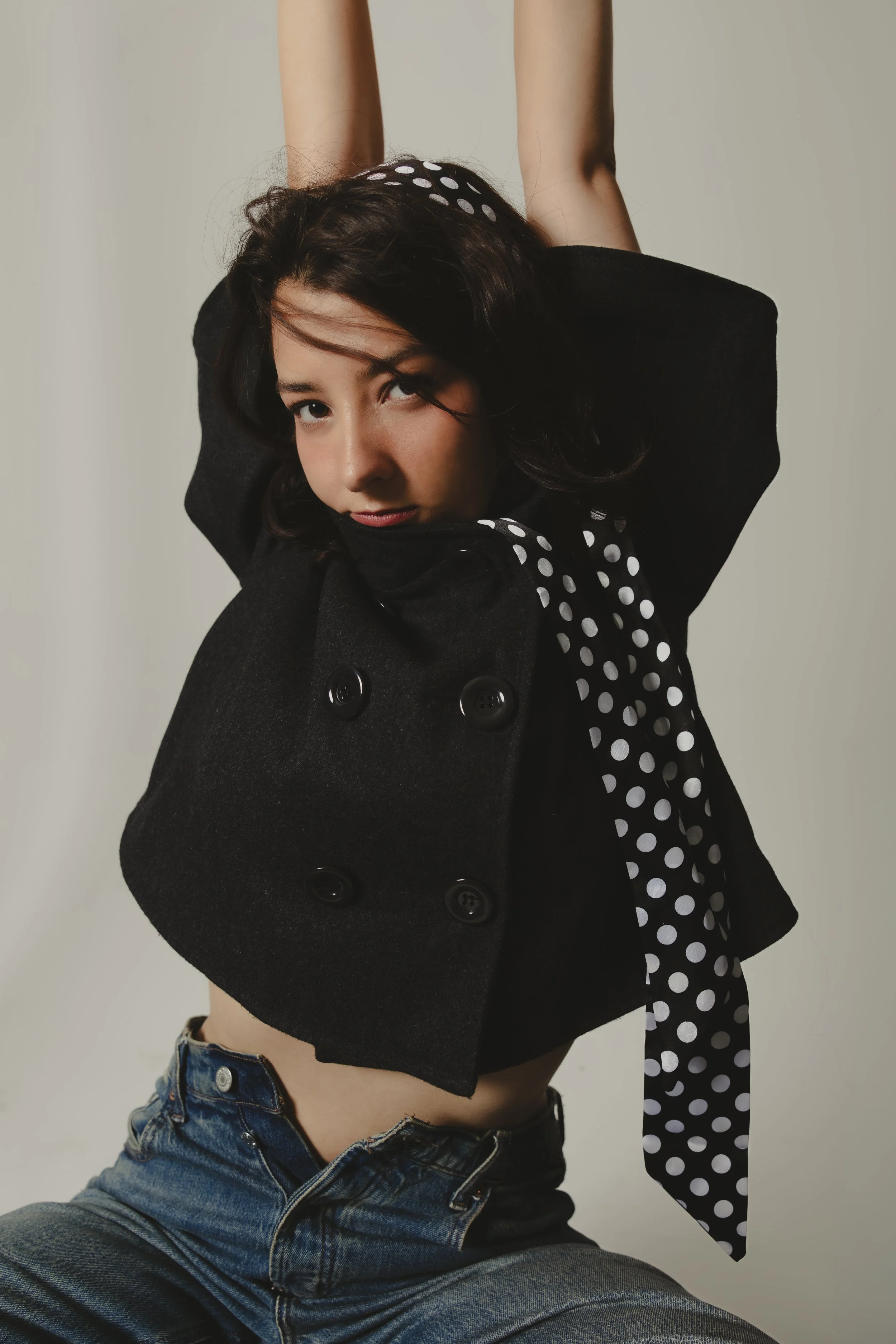 A young woman with dark hair, wearing a black coat with large buttons, a polka dot scarf, and jeans, sitting with her legs apart, with her arms raised above her head, against a plain background.