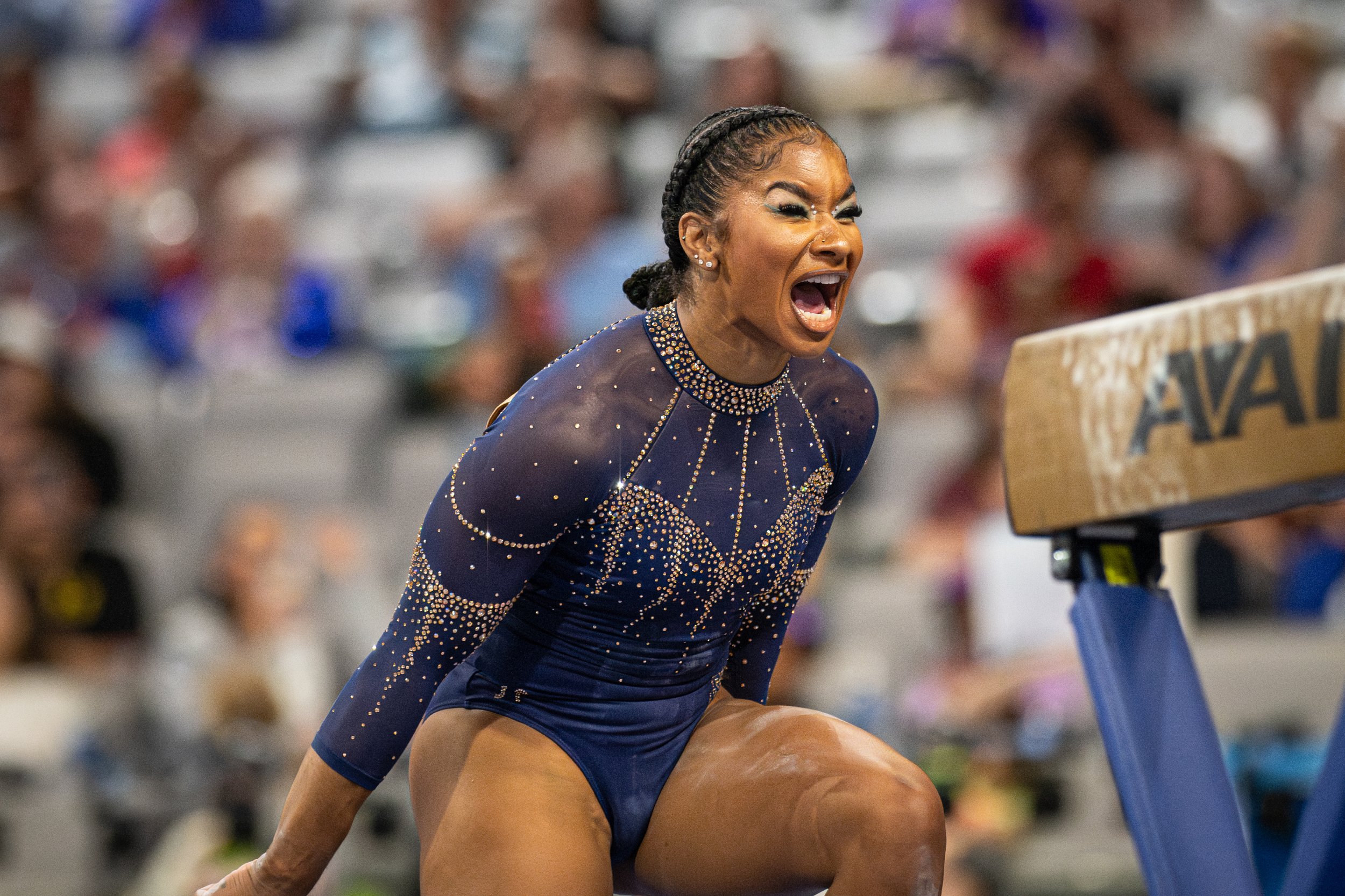 A female athlete, dressed in a blue and gold uniform with rhinestone decorations, is competing in a track and field event. She is shouting in a moment of intense effort or celebration, with a crowd of spectators blurred in the background.
