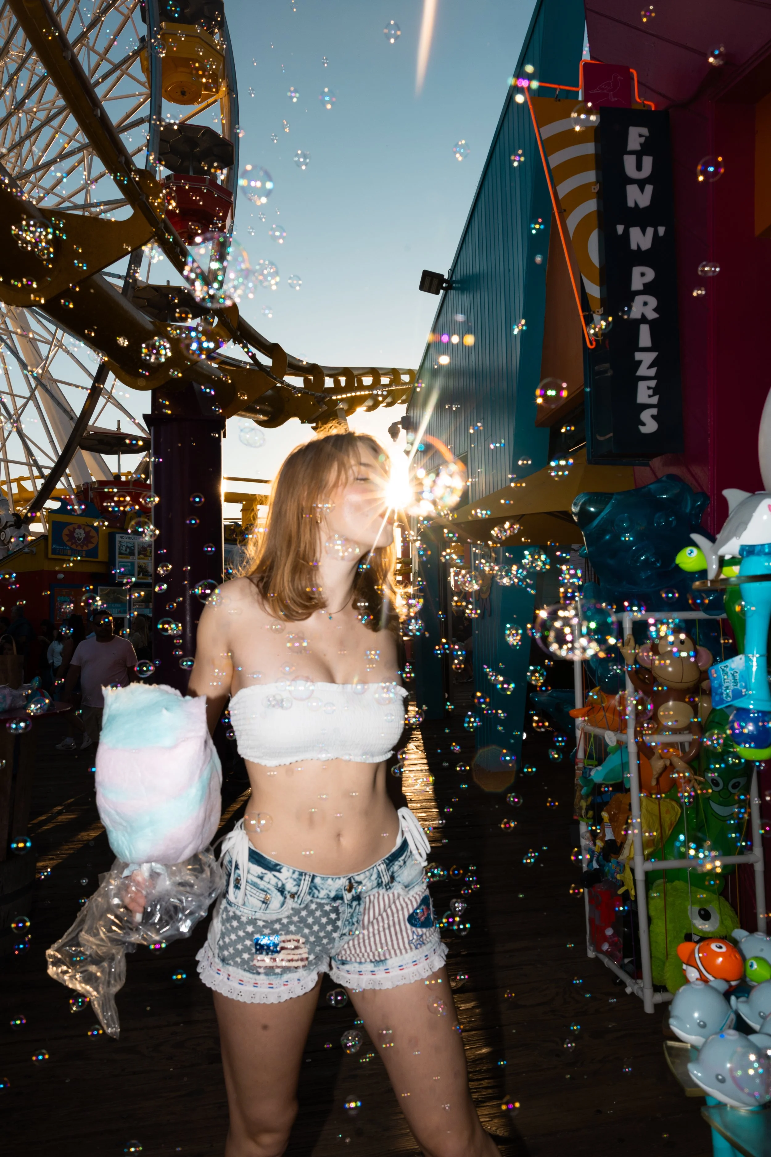 A woman in a tube top and shorts standing at a carnival with a Ferris wheel in the background. She is surrounded by floating bubbles and holding a large cotton candy.