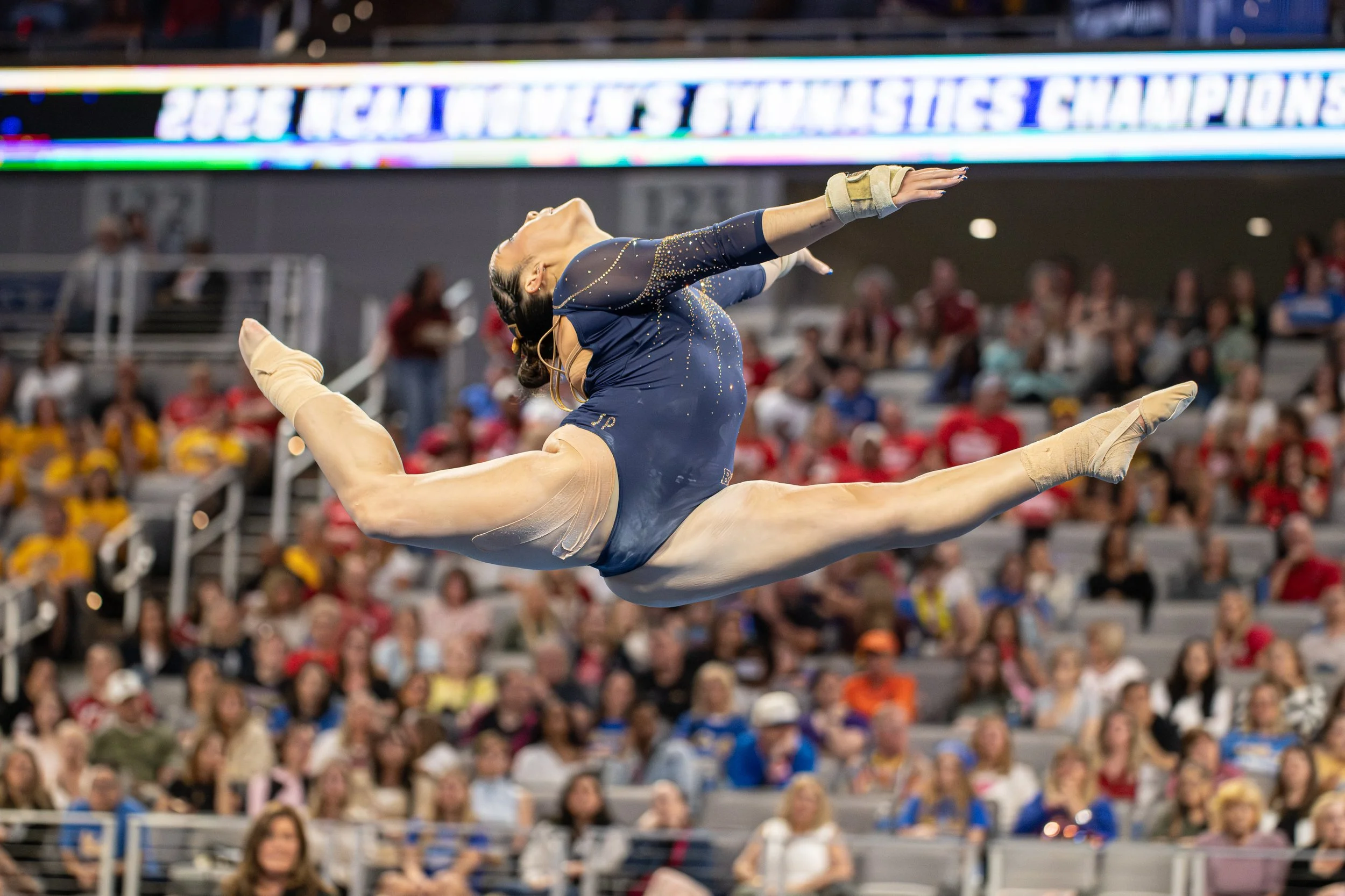 Female gymnast performing a leap during a competition with an audience in the background.