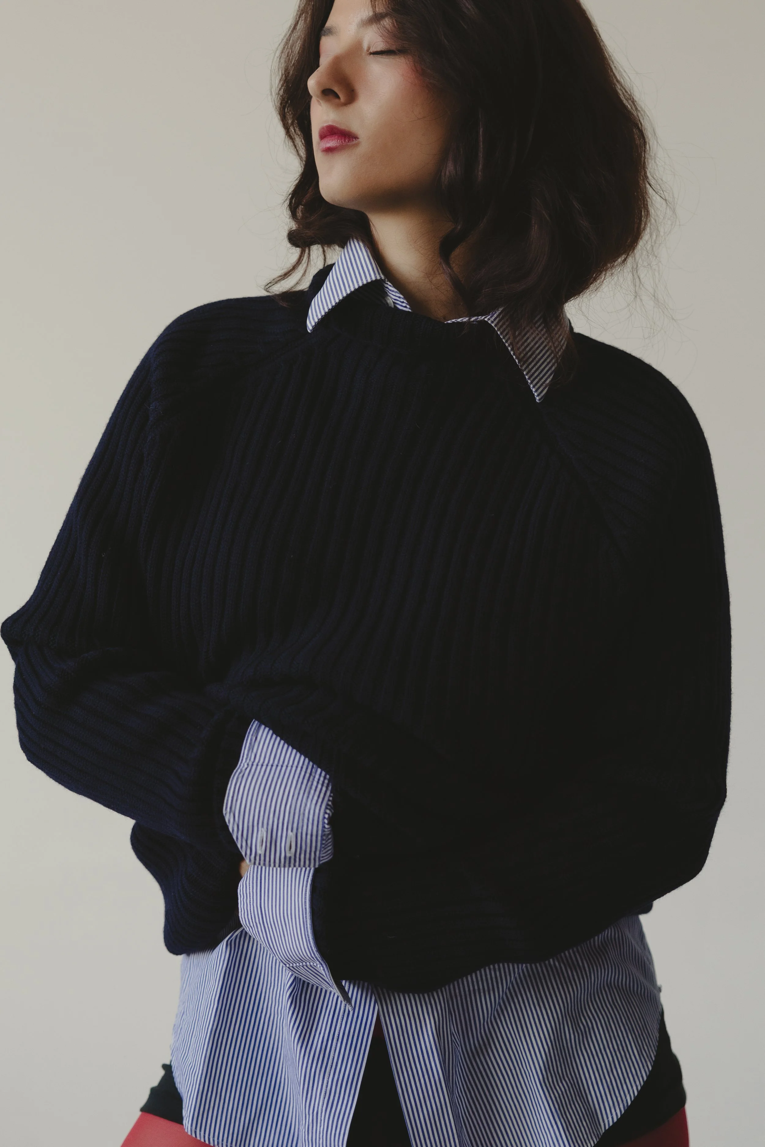 A woman with shoulder-length dark hair, wearing a black sweater over a blue and white striped shirt, standing against a plain background with her eyes closed.