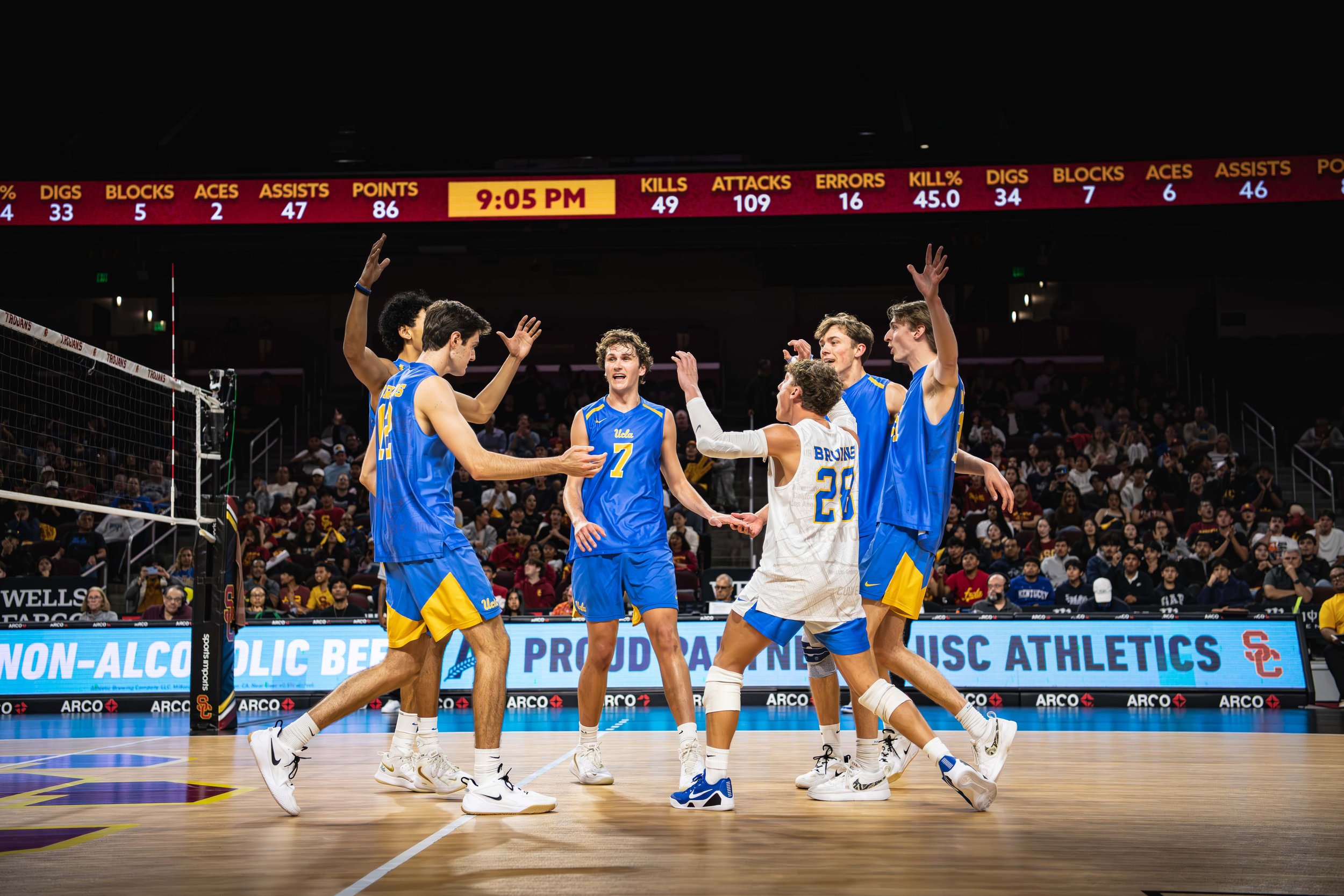 A group of UCLA volleyball players celebrating on the court during a match, with a scoreboard above them showing game stats and the time 9:05 PM.