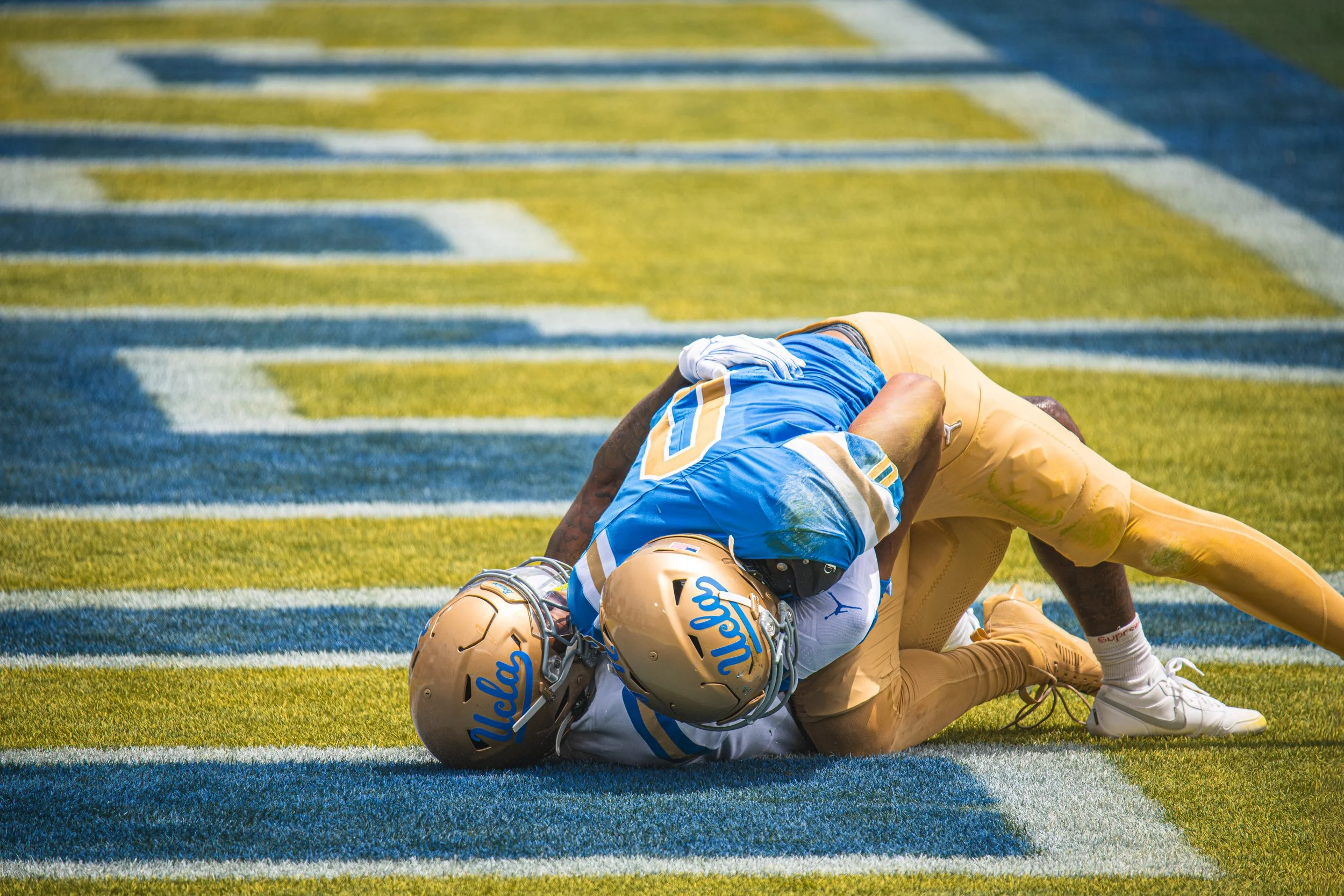 Two football players from UCLA and USC on the ground during a game, with UCLA's player in a white jersey and USC's player in a blue jersey, both wearing helmets and pads, on a football field with yellow and white yard lines.