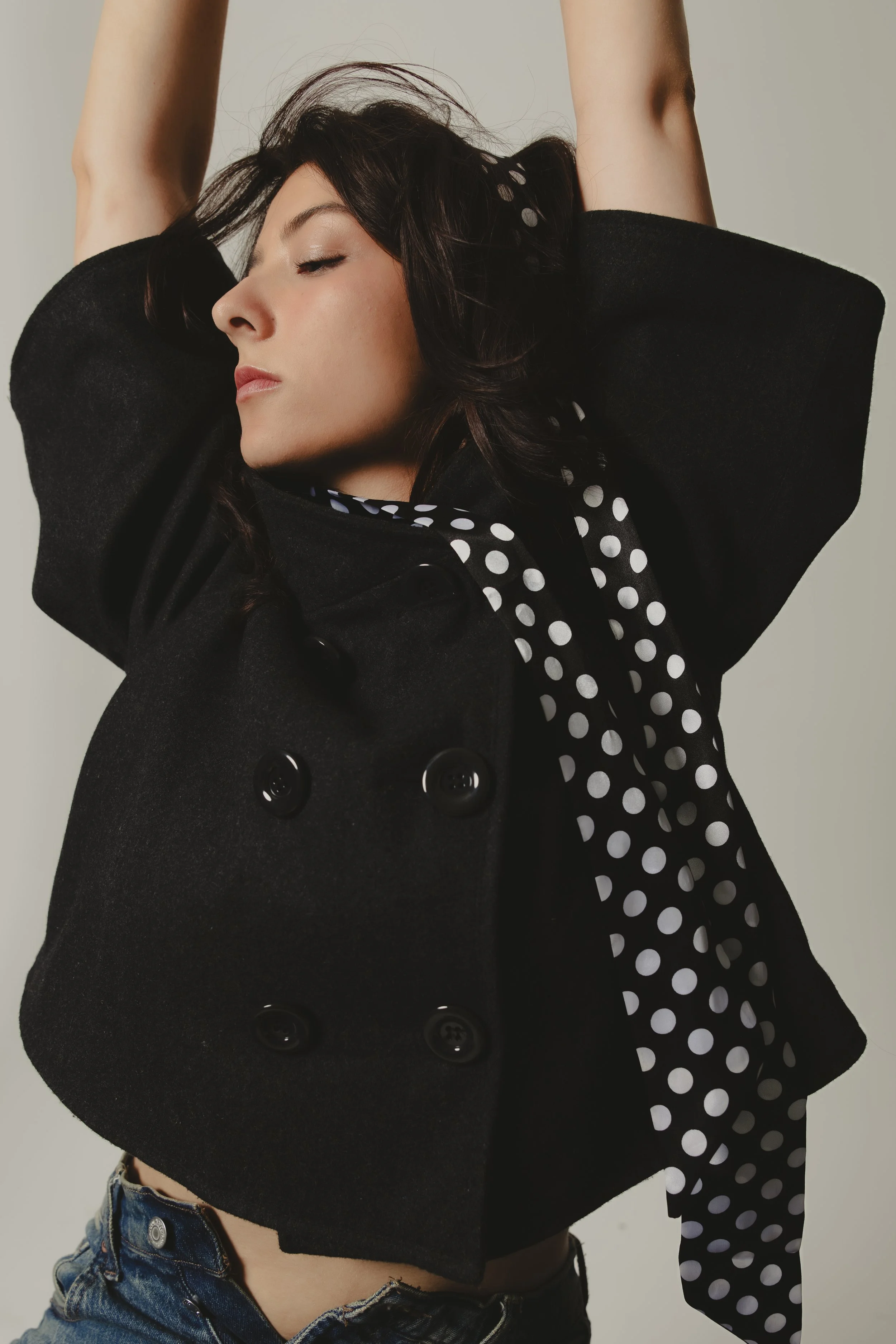A woman in a black double-breasted coat with black buttons and a polka-dotted scarf, wearing denim shorts. She has her arms raised above her head with her eyes closed, and her dark hair is slightly tousled.