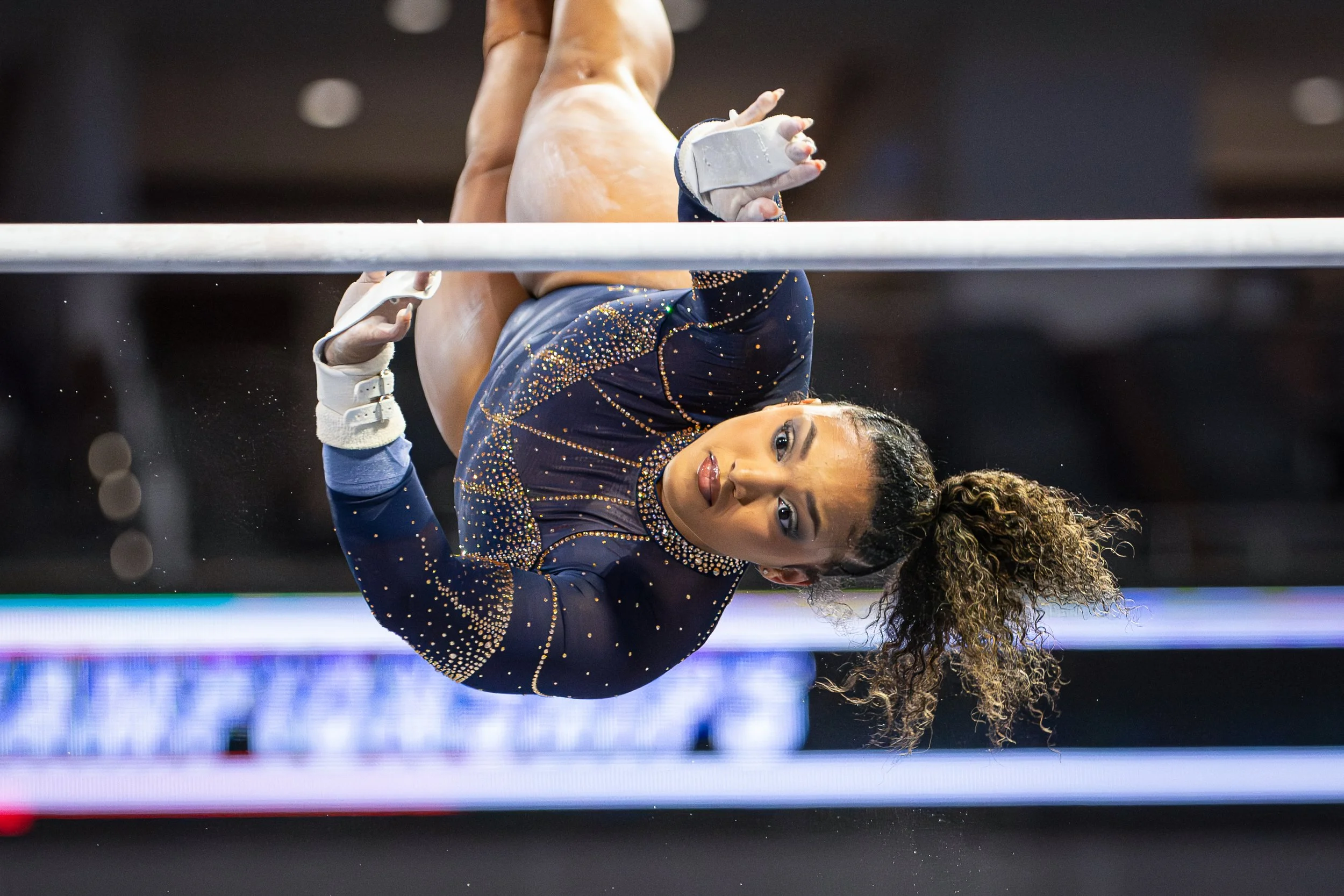 A female gymnast is performing an upside-down move on the uneven bars during a gymnastics competition. She is wearing a navy blue leotard decorated with gold and silver rhinestones and has curly hair.