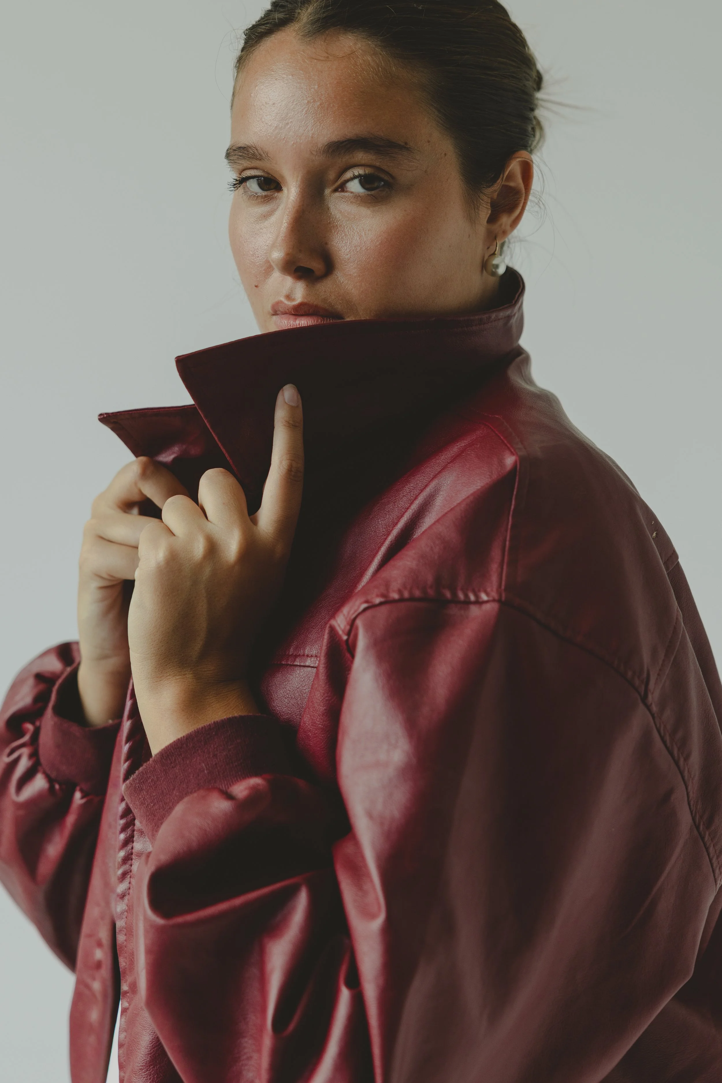 A woman with dark hair pulled back, wearing earrings and a red leather jacket, holding the collar of the jacket close to her face, with a neutral expression, against a plain light background.