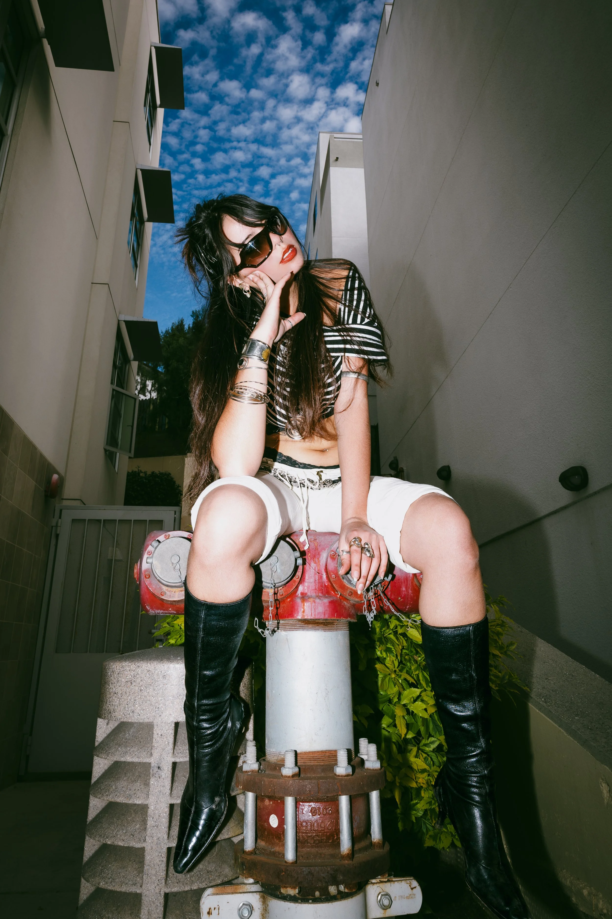 A young woman with long dark hair, wearing sunglasses, red lipstick, a black and white striped top, white shorts, and thigh-high black boots, is sitting on an artistic object that resembles a fire hydrant, outdoors against the background of a modern 