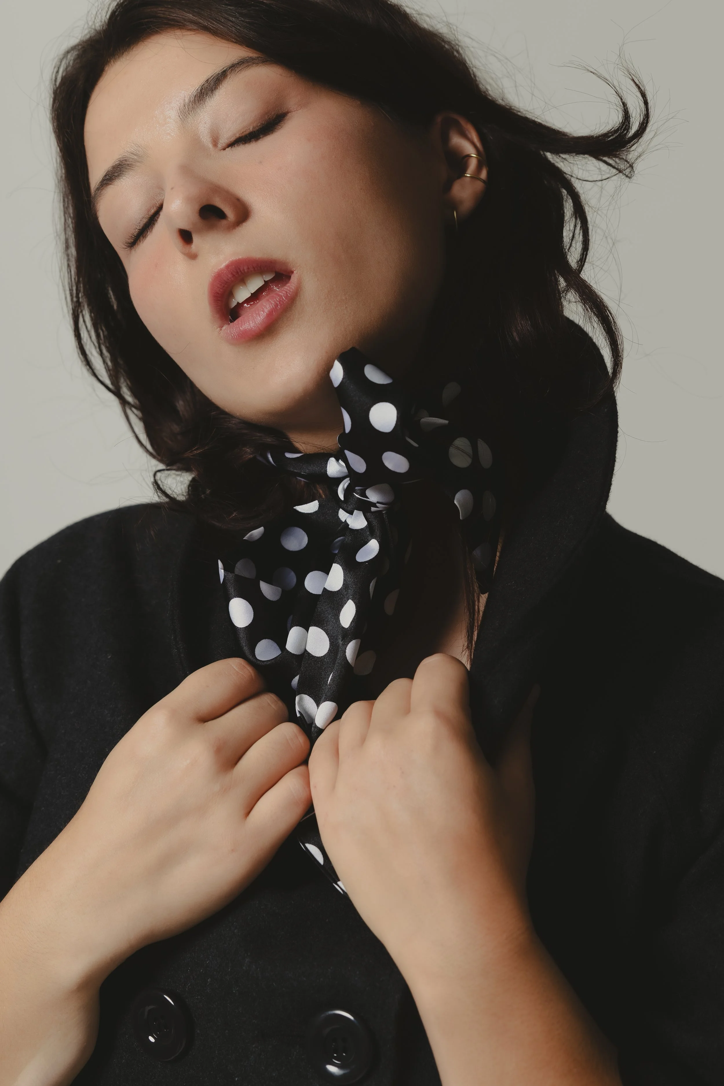 A woman with dark hair, jewelry in her ear, wearing a black outfit with large black buttons, holding a black and white polka-dot scarf near her face, eyes closed, head tilted back.