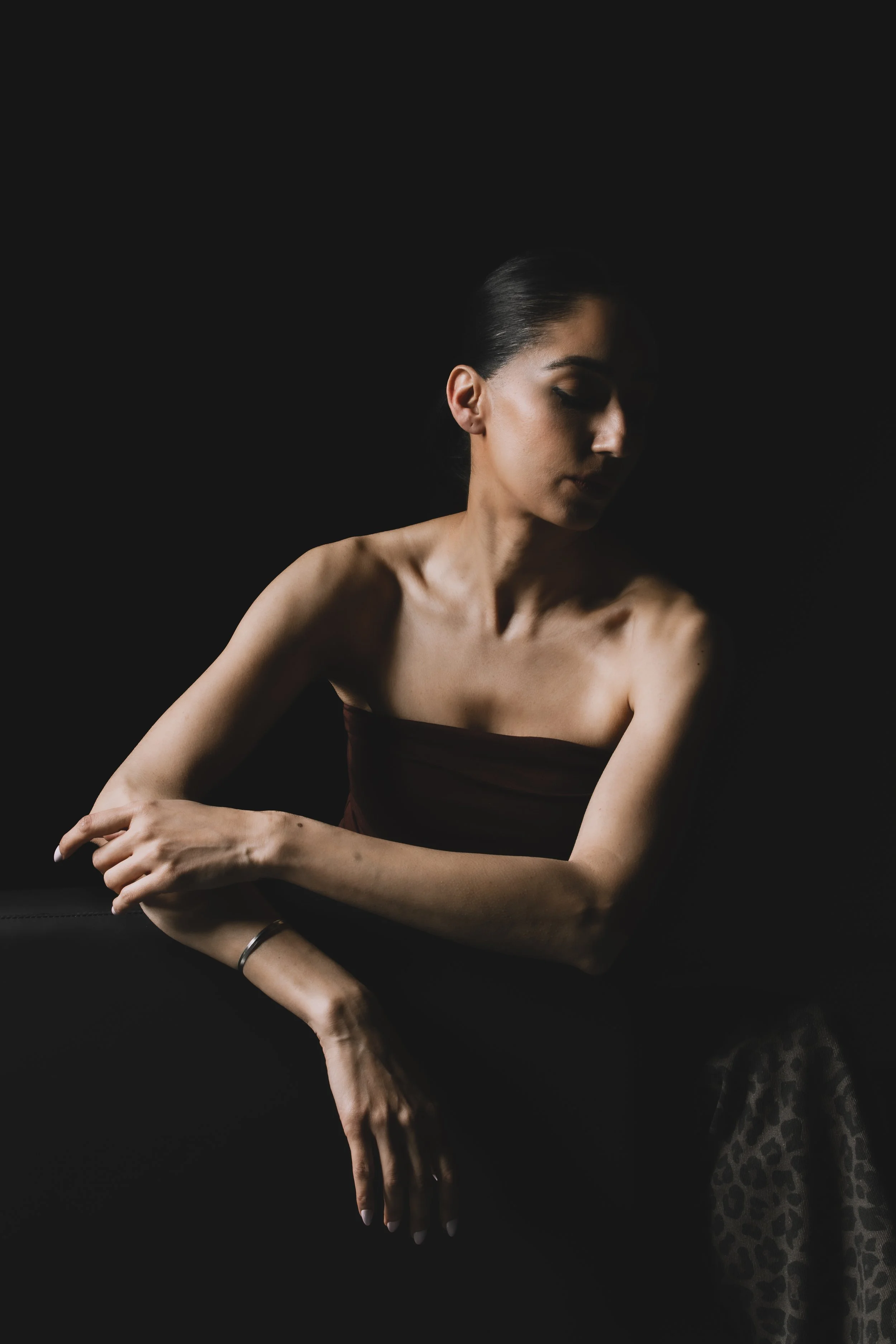 A young woman with dark hair tied back, wearing minimal makeup, poses against a black background with her eyes closed, her left arm crossed over her body, and her right arm resting on her left arm. She is wearing a strapless top and leopard print pan