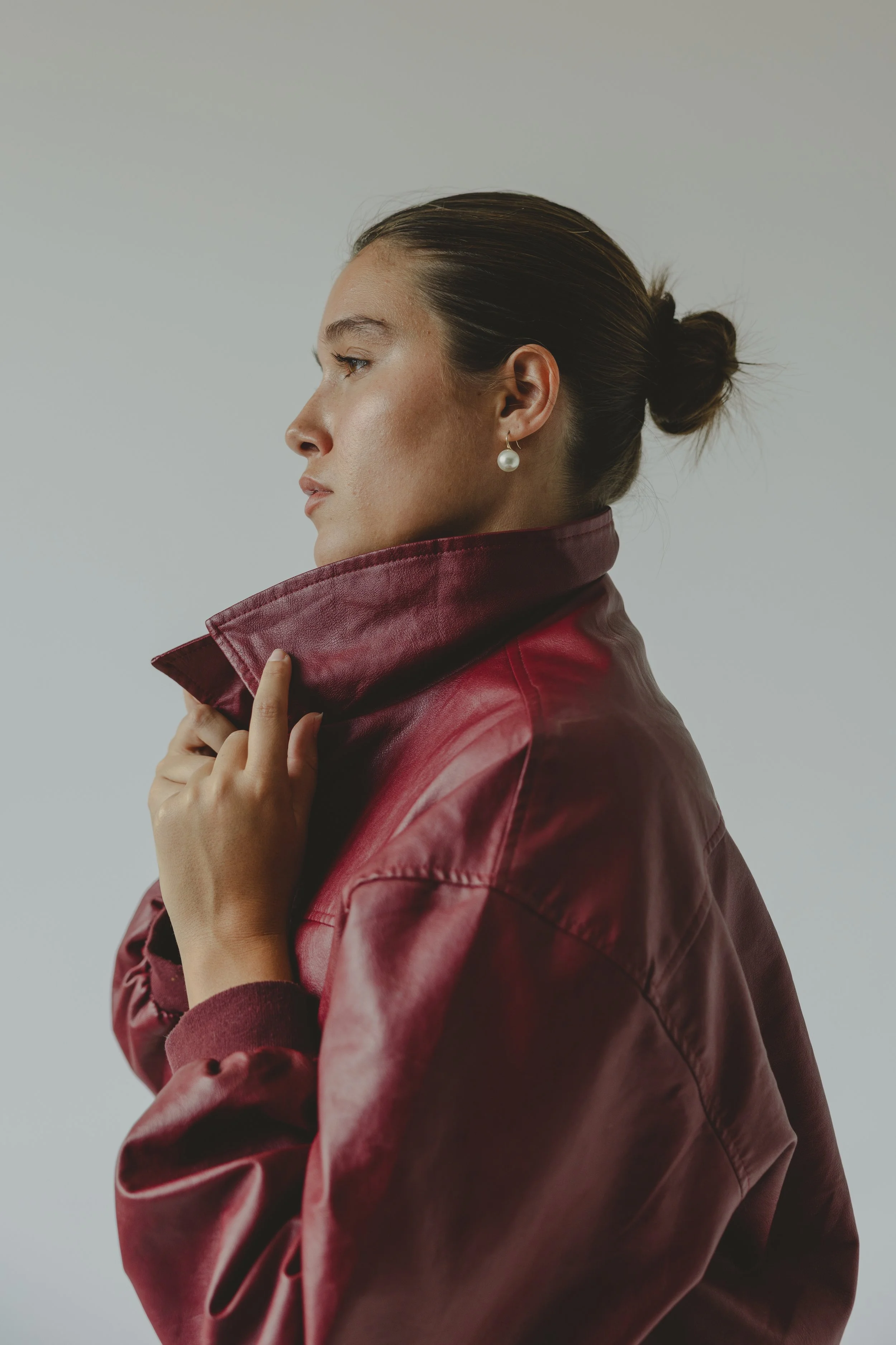 Side view of a woman with brown hair tied in a bun, wearing pearl earrings and a red leather jacket with the collar turned up against a plain background.