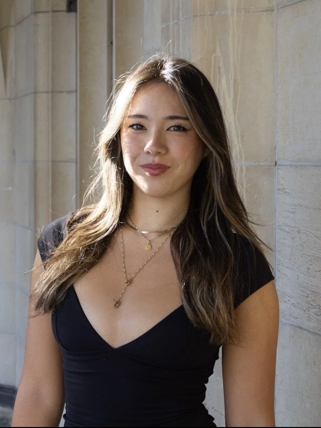 Young woman with long brown hair and light complexion smiling outdoors against a stone wall, wearing a black top and layered gold necklaces.