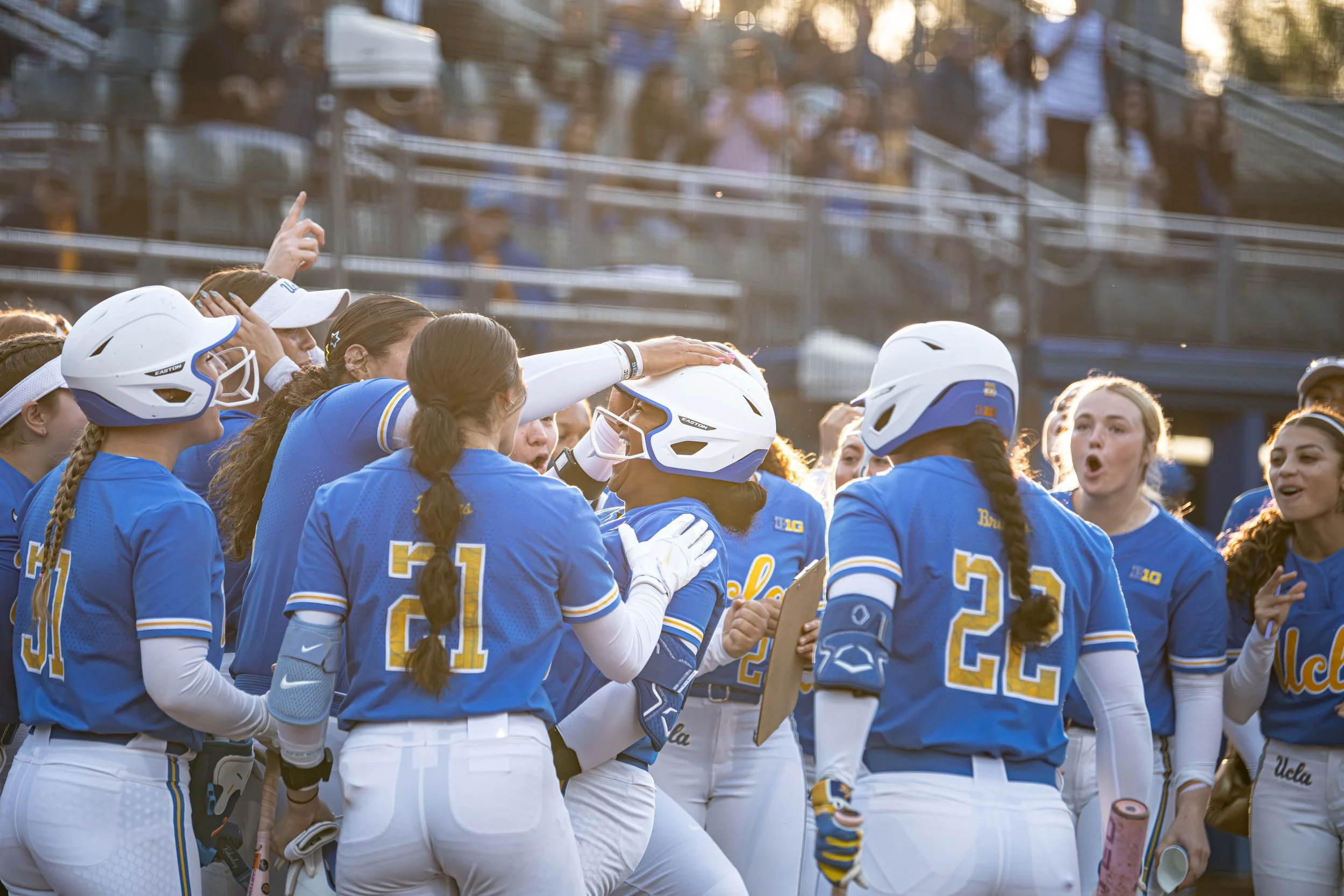 A group of female softball players in blue and white uniforms gathered together, some wearing helmets, engaging in a team discussion or celebration on the field.