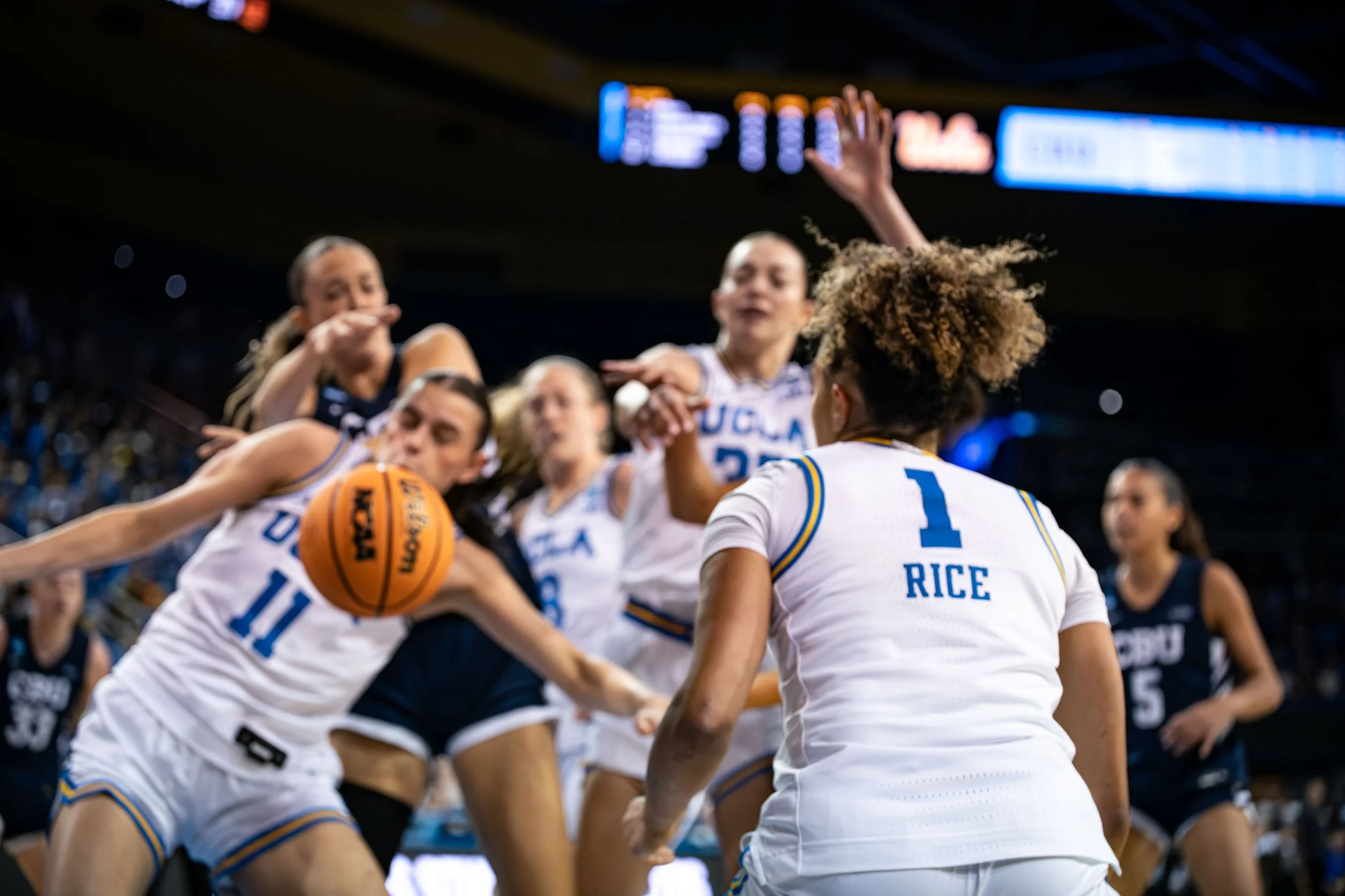 UCLA women's basketball team huddles during a game, with players in white jerseys with blue and yellow accents, on the court with a basketball in play.